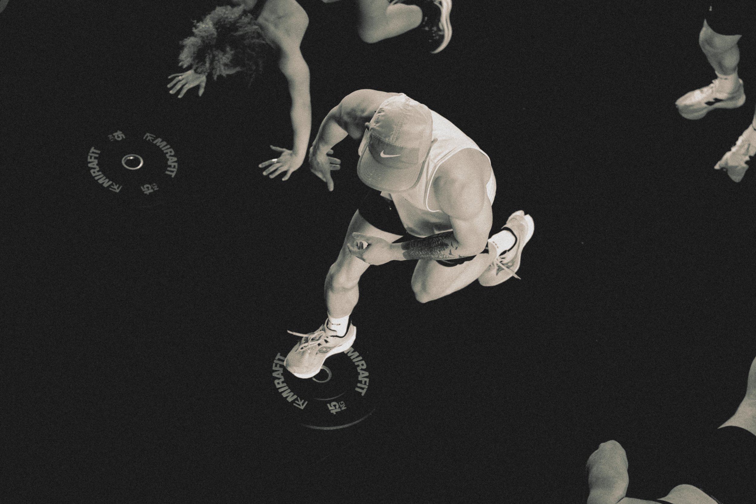 A group of people participating in a fitness class, performing exercises on a dark gym floor with a fitness circle in the center. The focus is on a man wearing a hat, tank top, shorts, and sneakers, crouching on one knee while stretching or preparing to exercise. Other individuals are visible in the background, also engaged in physical activity.