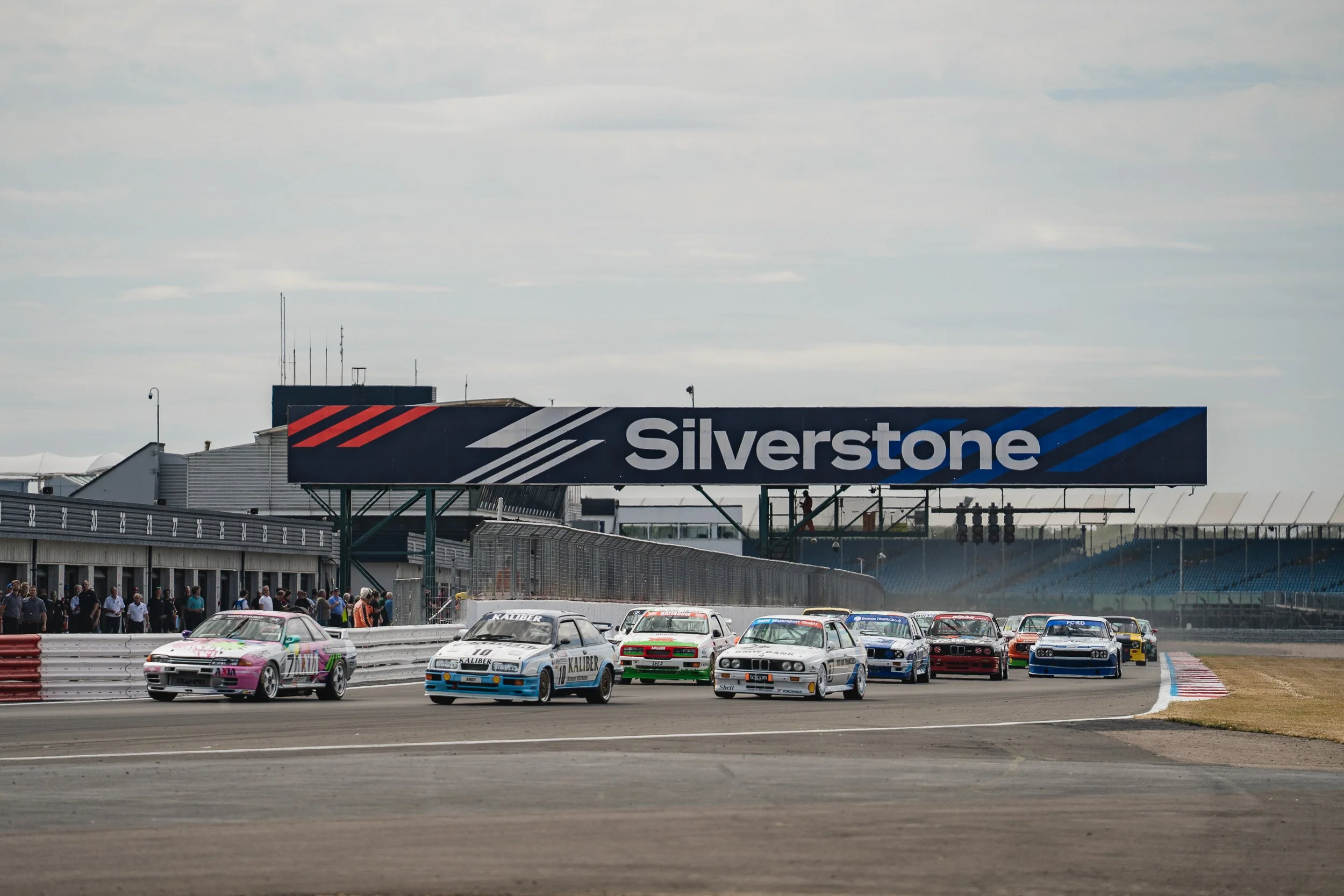 Pack of touring cars racing side by side under the Silverstone gantry at the BRDC Classic