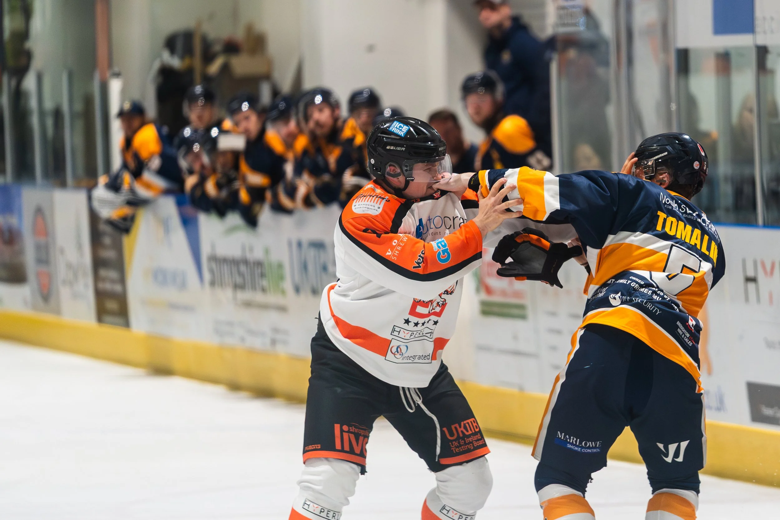 Two hockey players fighting on the ice rink during a game with teammates on the bench in the background.