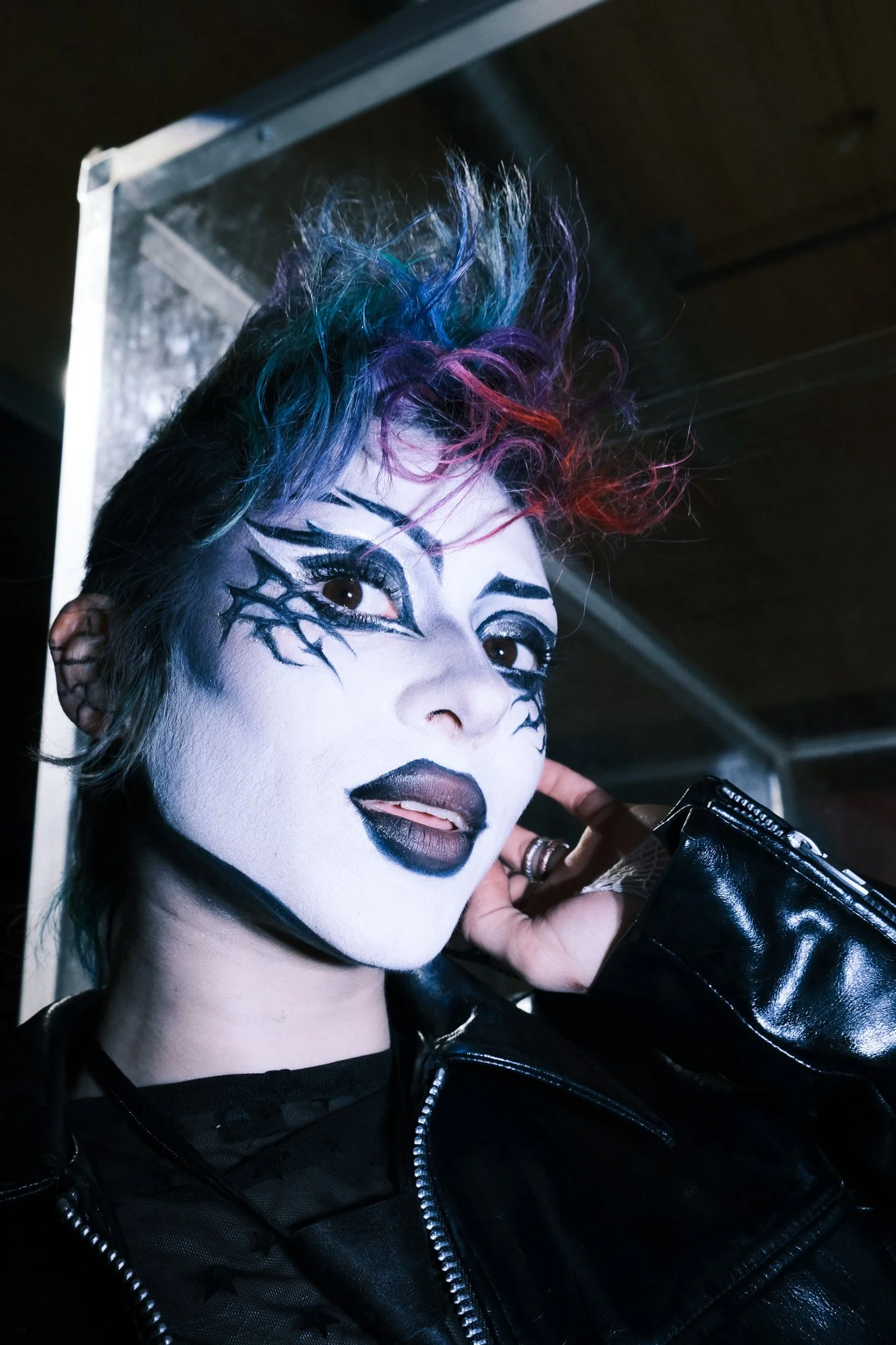 A person with face paint resembling a spooky clown or demon, wearing a black leather jacket, with colorful hair styled in a spiky mohawk, and holding a phone to their ear.