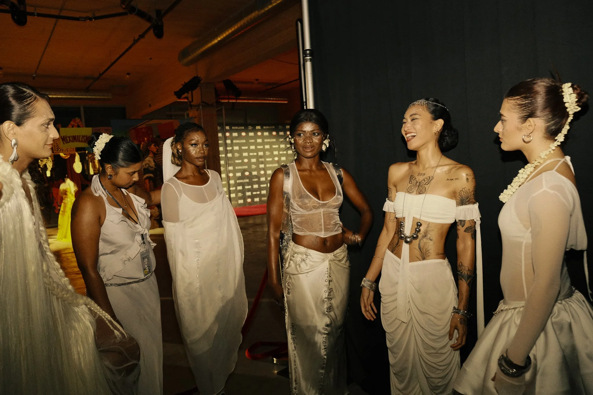 Group of women wearing traditional white dresses, standing and talking backstage at an event.