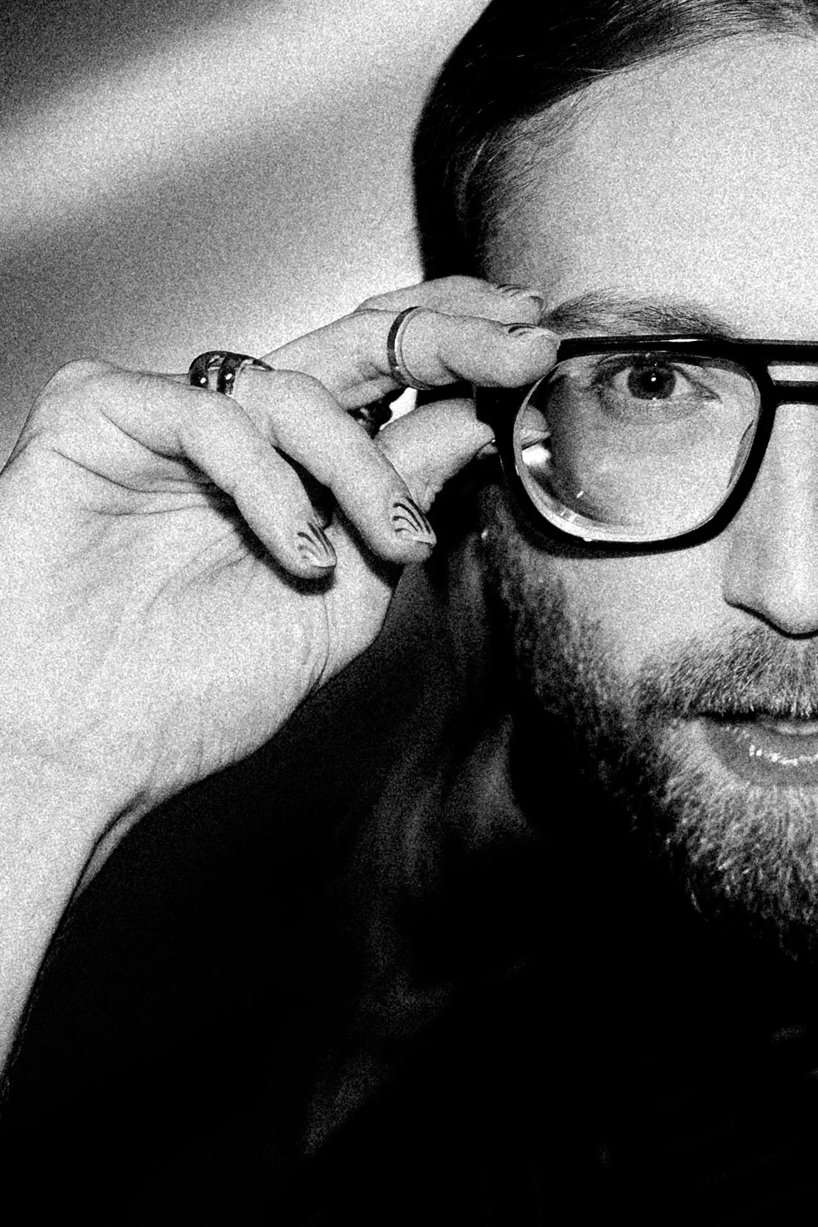 Close-up black and white photo of a man with a beard and glasses, pulling down his glasses with his hand, which has rings and painted nails, revealing his eye.