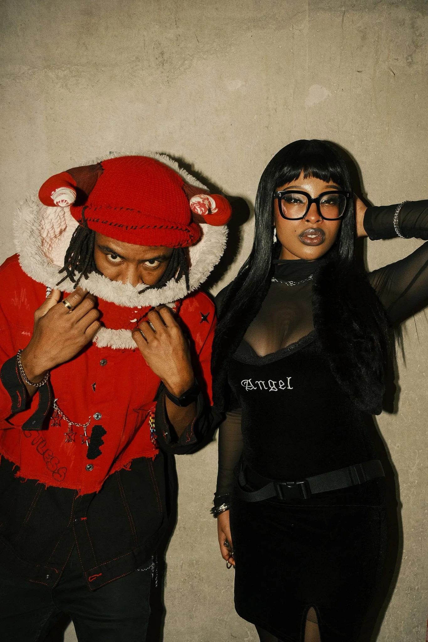 A person dressed in a red hat with a pom-pom and a red jacket, holding the collar, standing next to a woman wearing black glasses, a black sheer top with 'Angel' written on it, and a black skirt, against a plain wall.