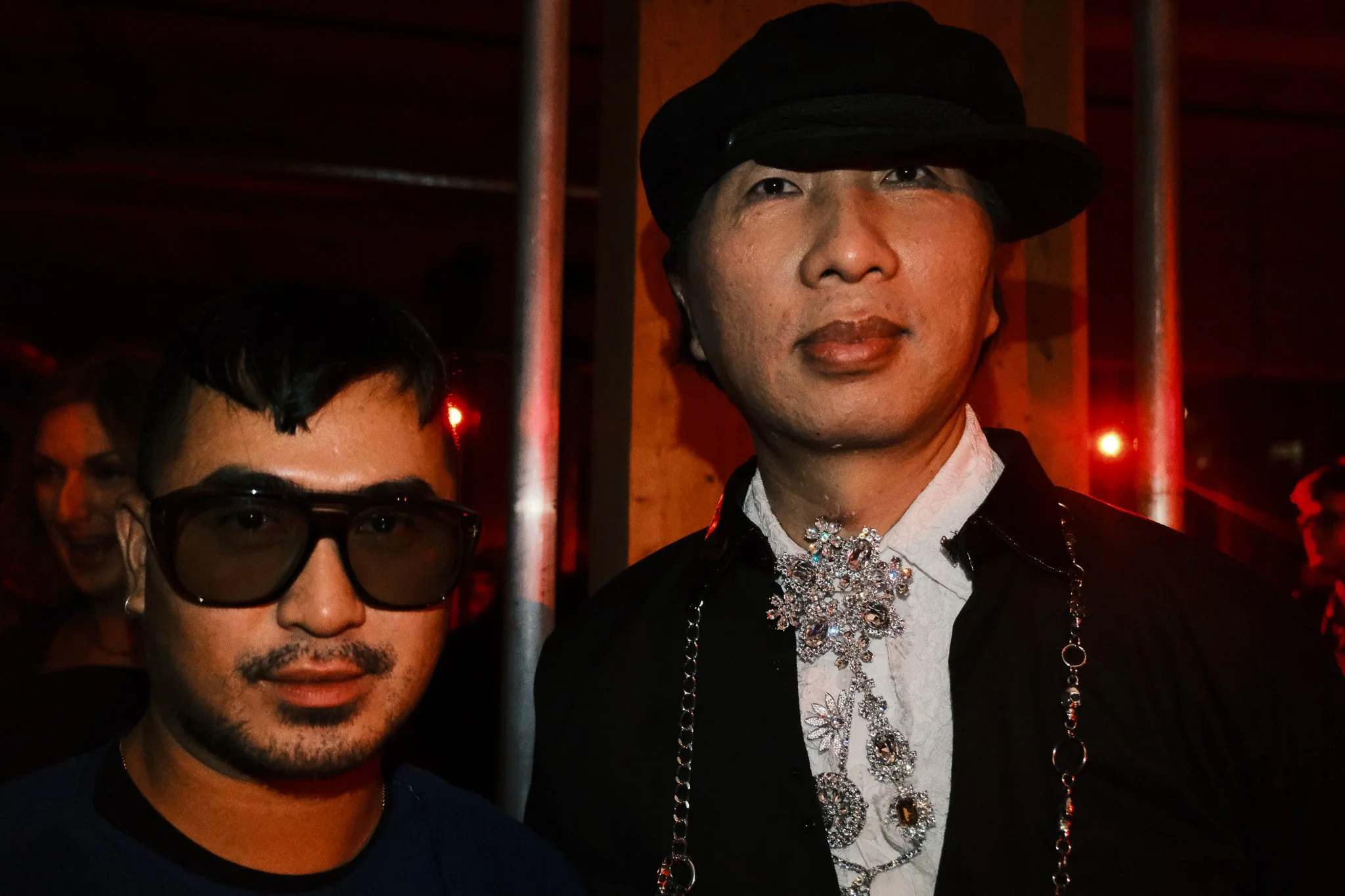 Two men at a social event, one wearing large glasses and the other wearing a black hat and jewelry, in a dimly lit environment with red lighting.