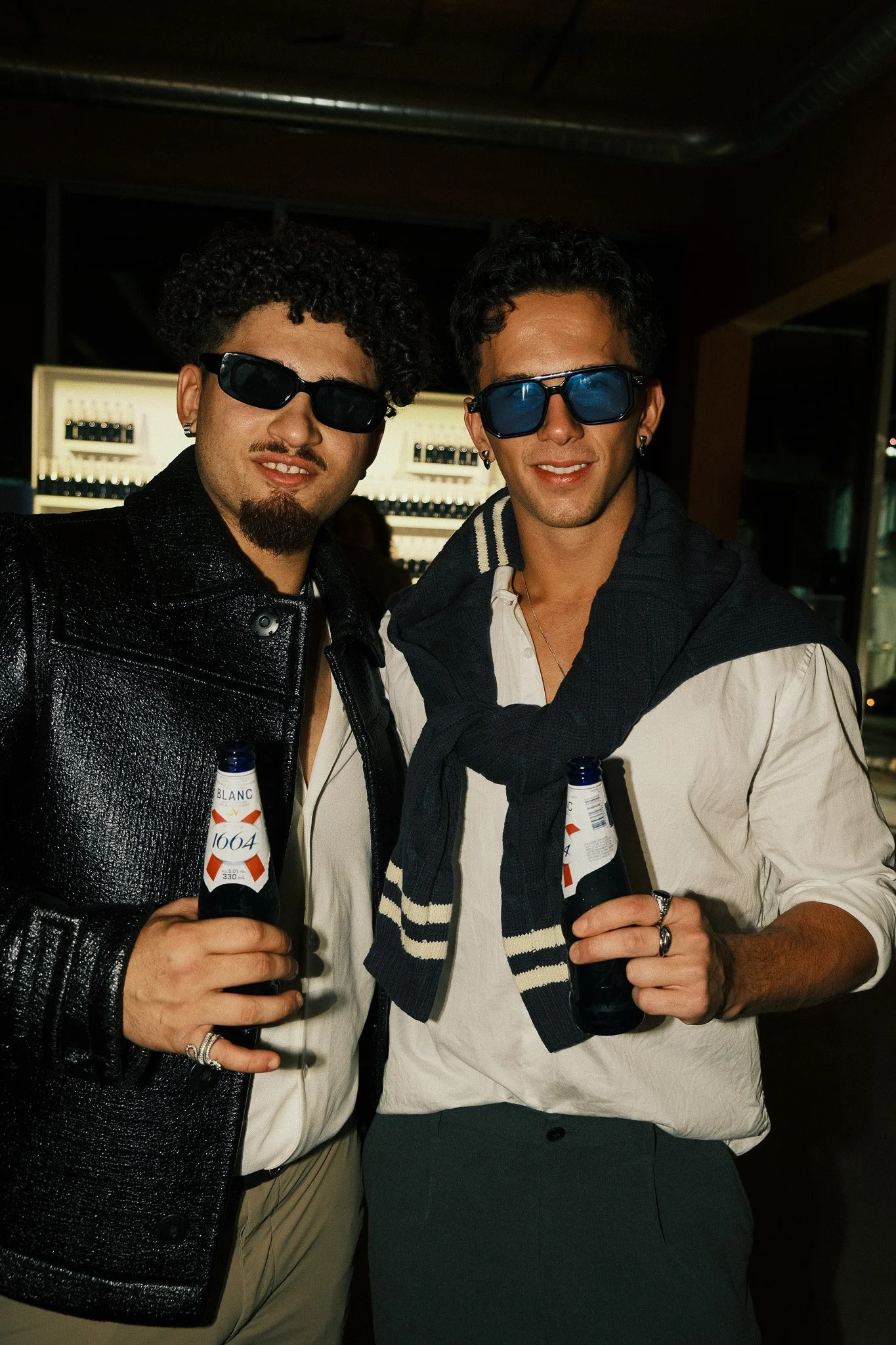 Two young men wearing sunglasses and holding beer bottles, standing close together indoors at a social event.