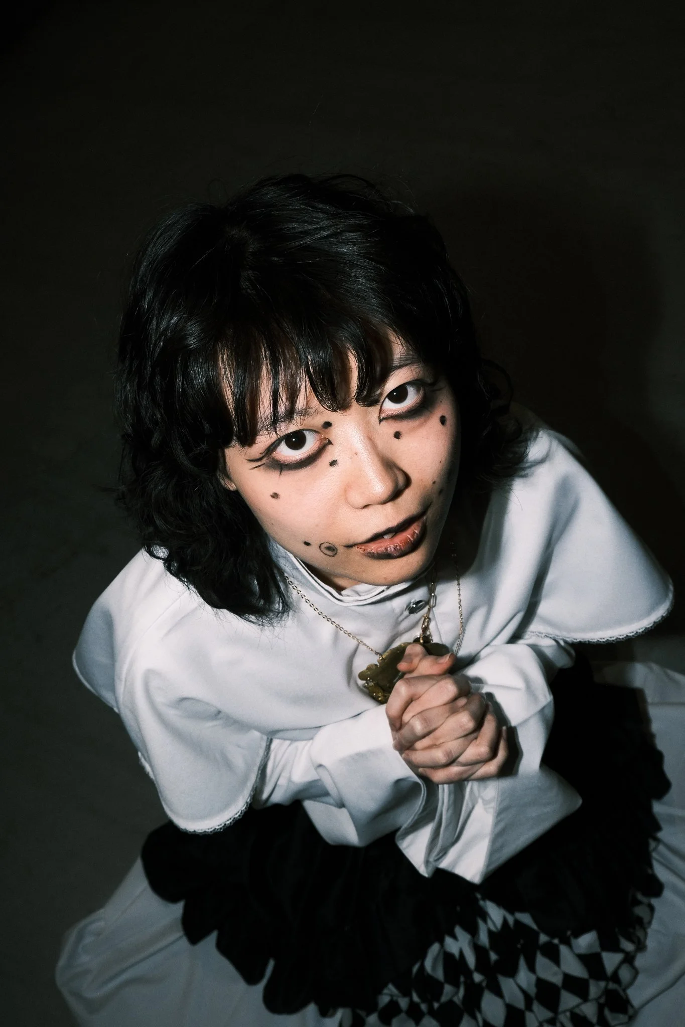 A person with black, wavy hair and dramatic makeup looks up at the camera. They are kneeling and holding their hands clasped together. They are wearing a white coat with lace trim and a necklace with a large pendant, against a dark background.