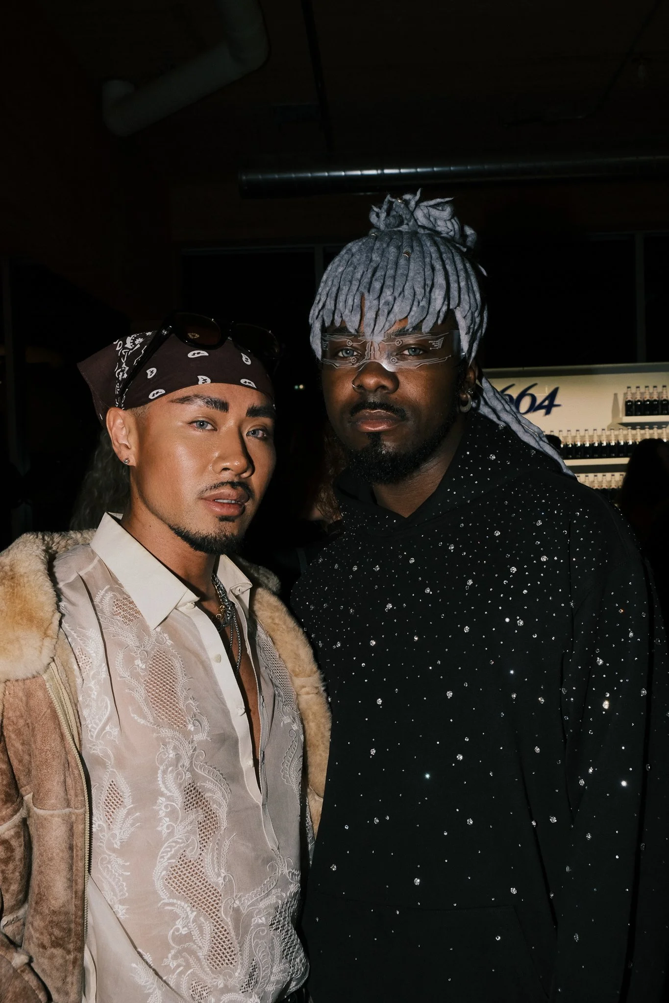 Two men standing close together in a dimly lit setting, one with a bandana and the other with gray dreadlocks and stylish glasses, both dressed fashionably.