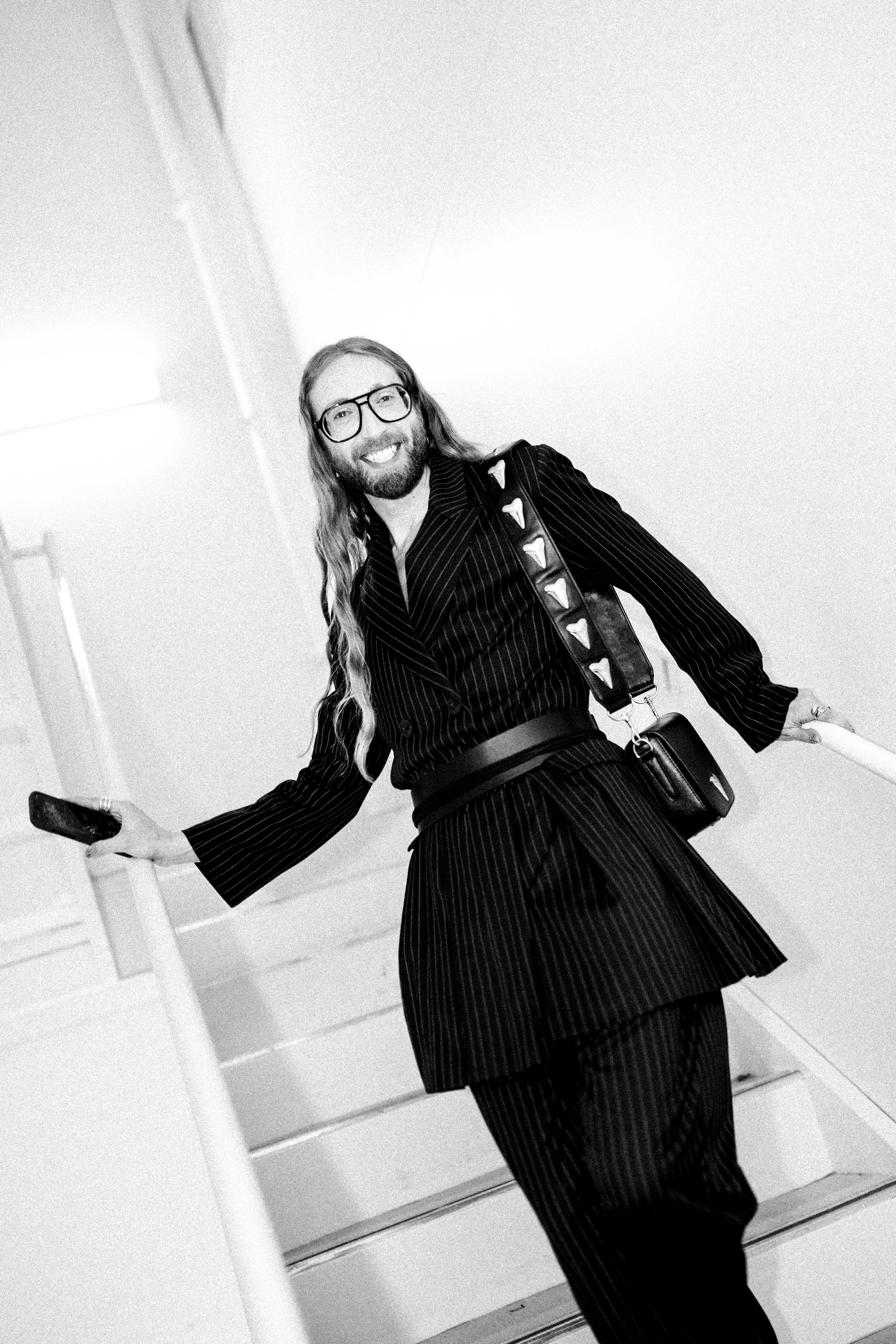 A man with long hair, glasses, and a beard smiling on a staircase, holding a phone in one hand and a railing with the other, wearing a pinstripe suit and a crossbody bag.
