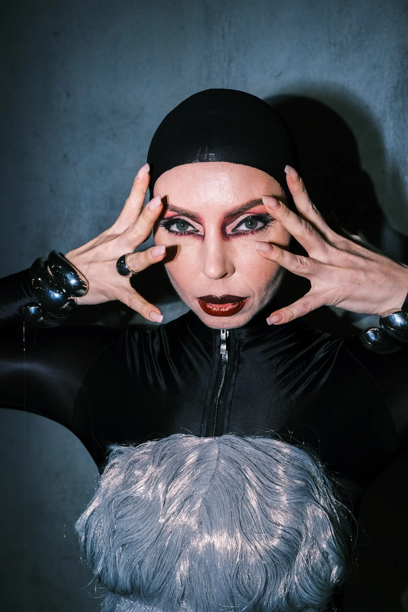 A drag queen with platinum blonde hair and dramatic makeup, wearing a shiny black bodysuit, holding her hands near her face, and looking directly into the camera.