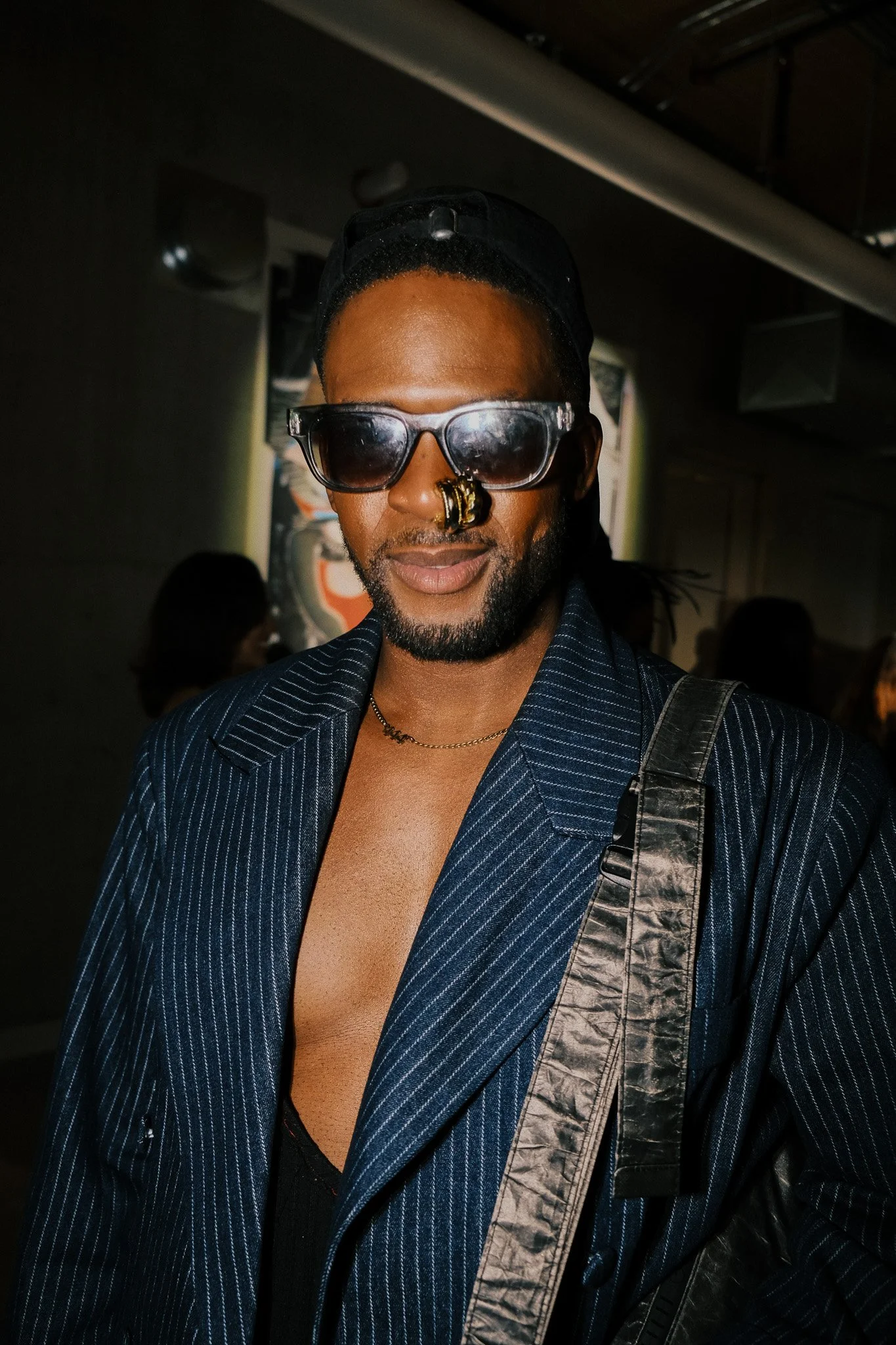 A man wearing sunglasses, a nose ring, and a dark blue pinstripe blazer with a black strap over his shoulder, in a dimly lit indoor setting