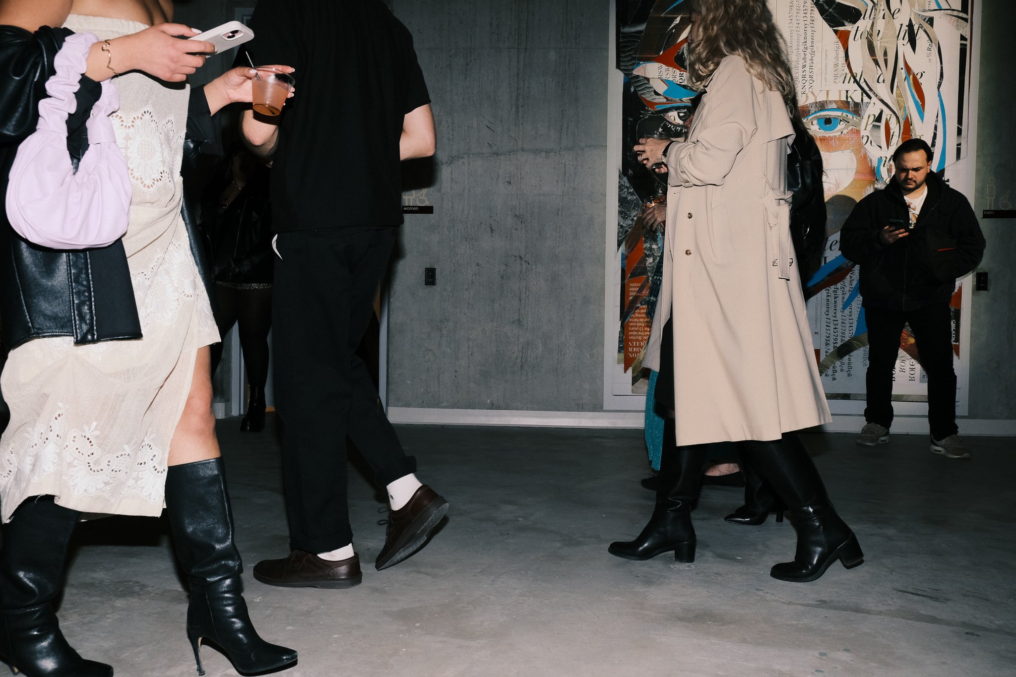 Group of people standing and walking indoors, some using their phones, wearing fashionable clothing including boots, dresses, and coats, in front of abstract artwork on a concrete wall.