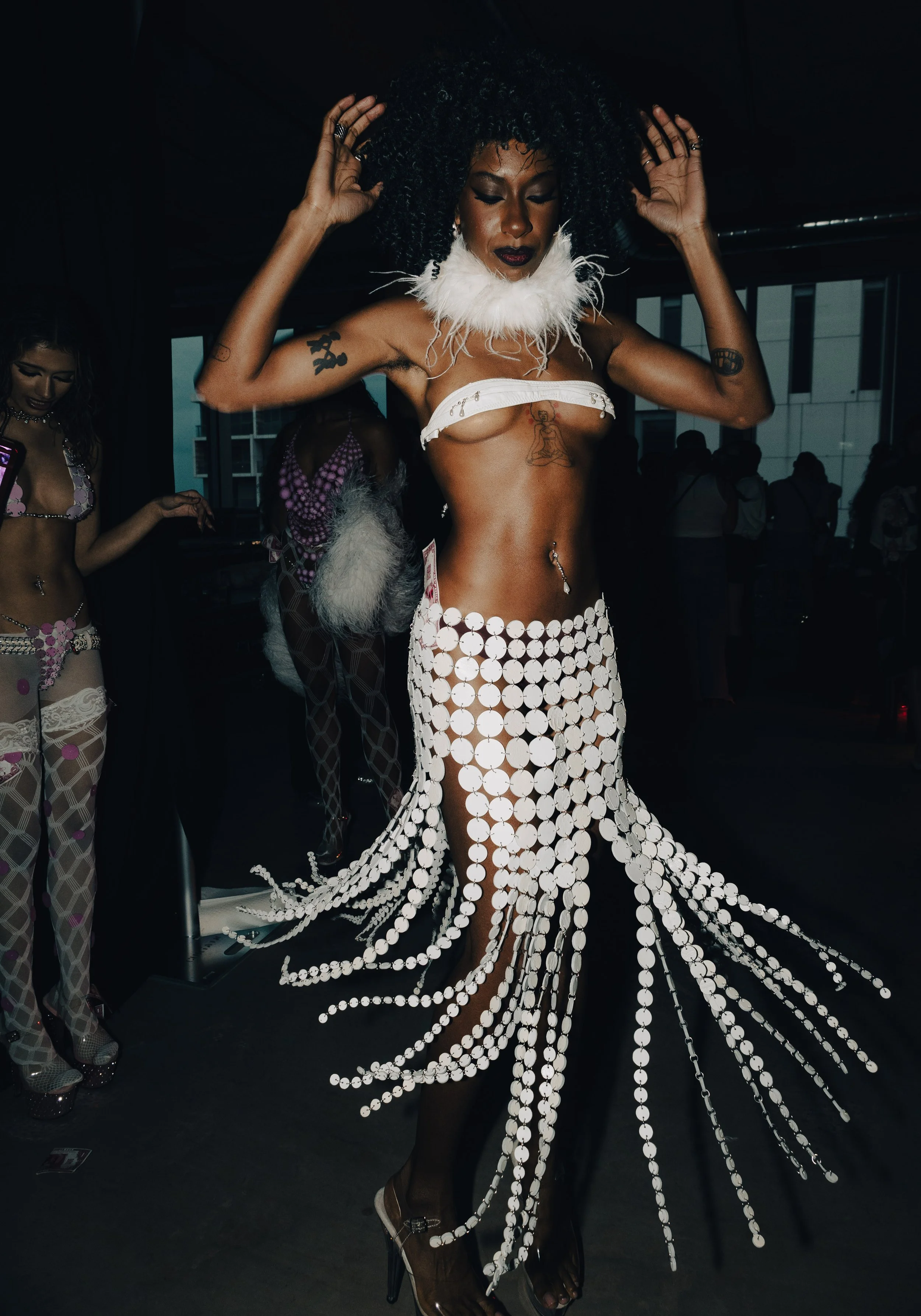 Woman wearing a white sea shell and feather costume with black tights and high heels, dancing or posing at an indoor event with other people in the background.