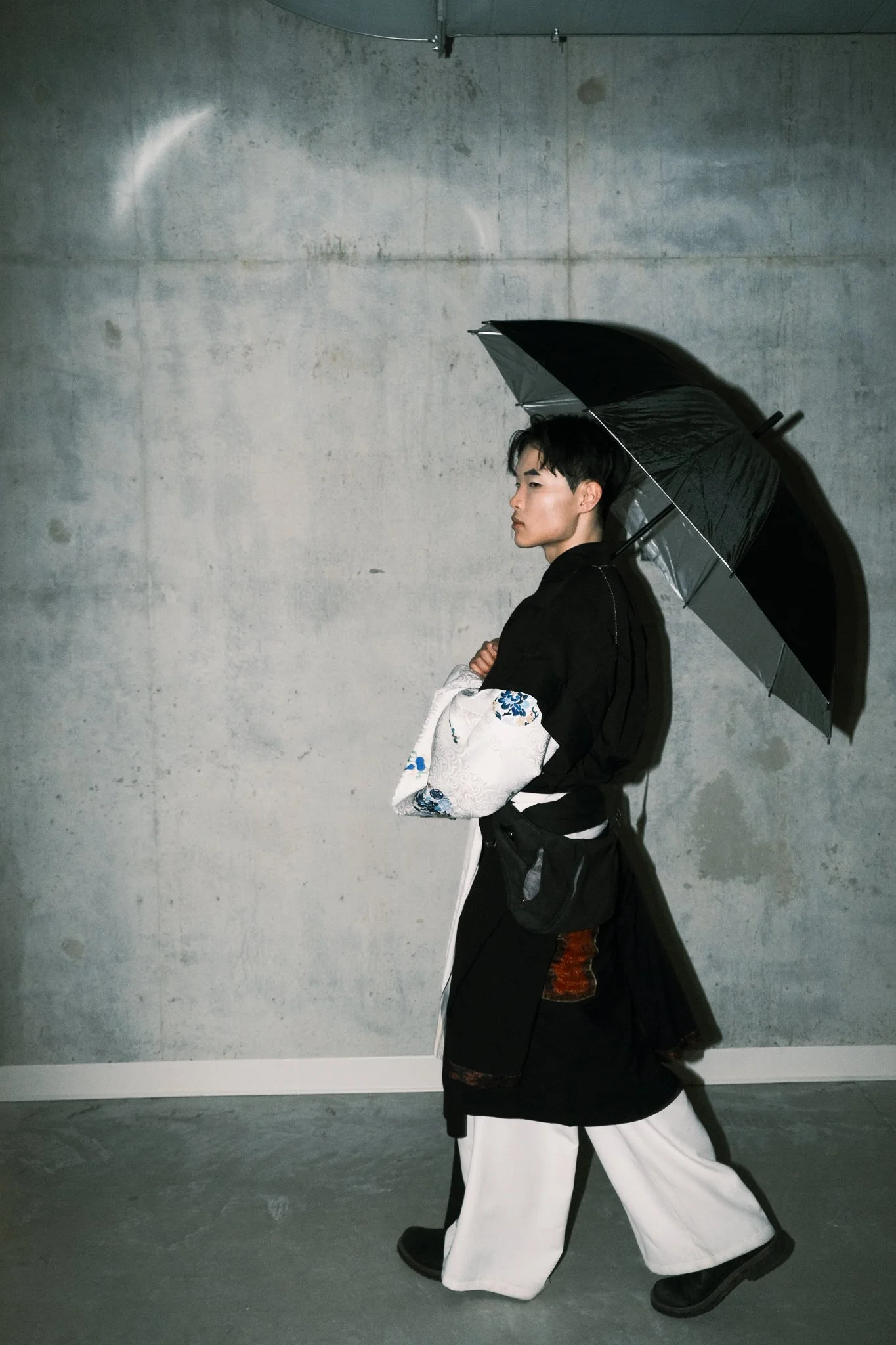 Young man walking with a black umbrella, wearing traditional and modern clothing, against a concrete wall.