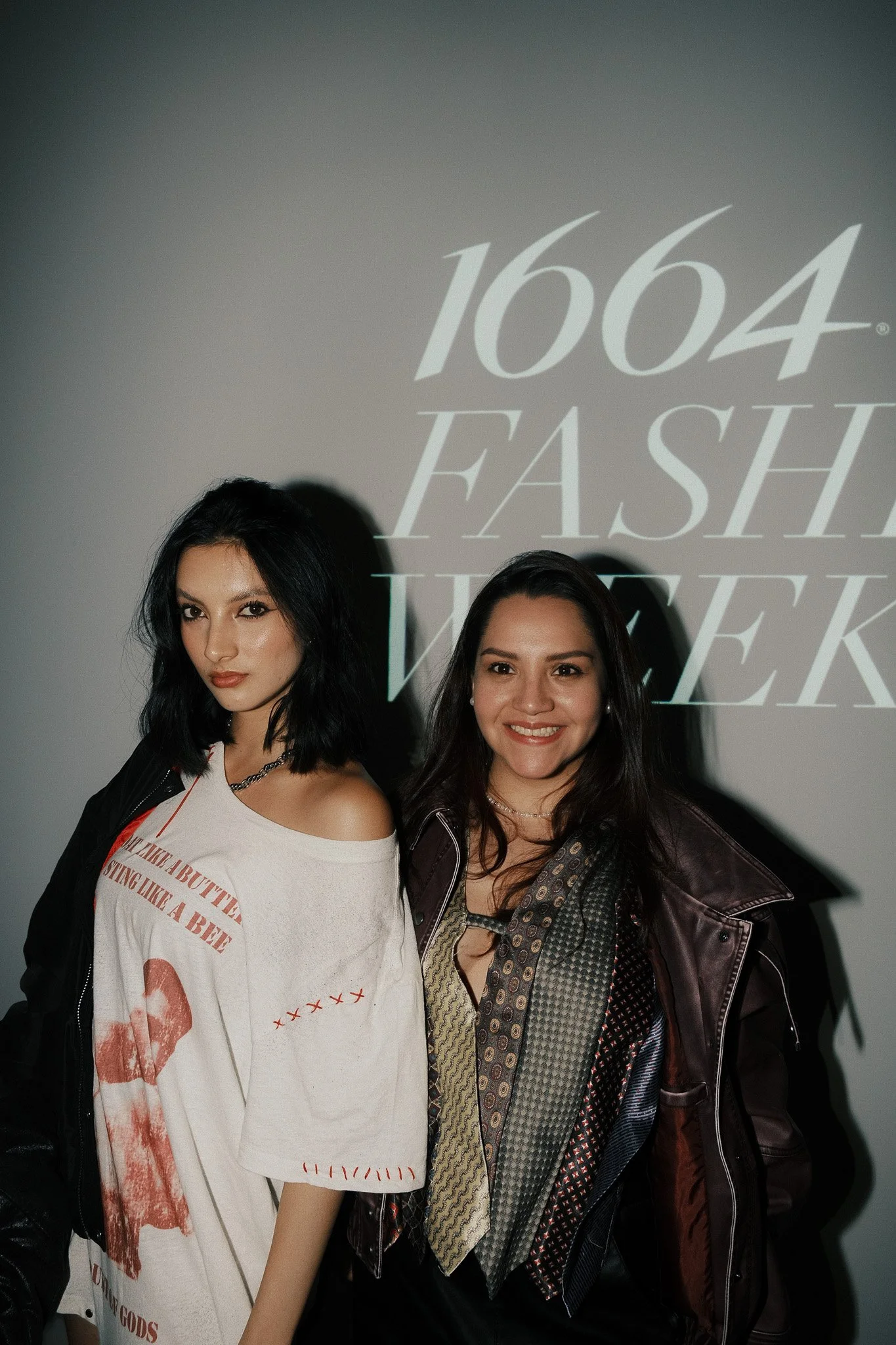 Two women pose in front of a projection that reads '1664 Fash Week'.