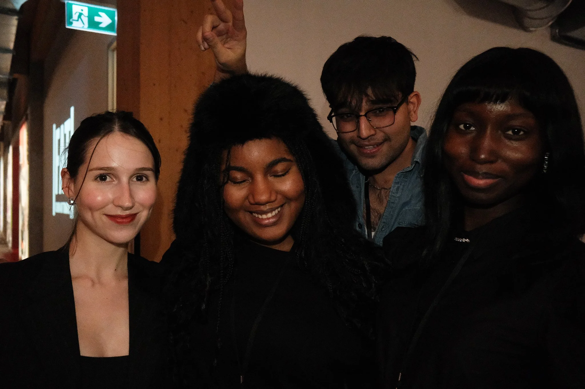 Four people close together indoors, smiling and posing for a photo, with a green exit sign visible in the background.