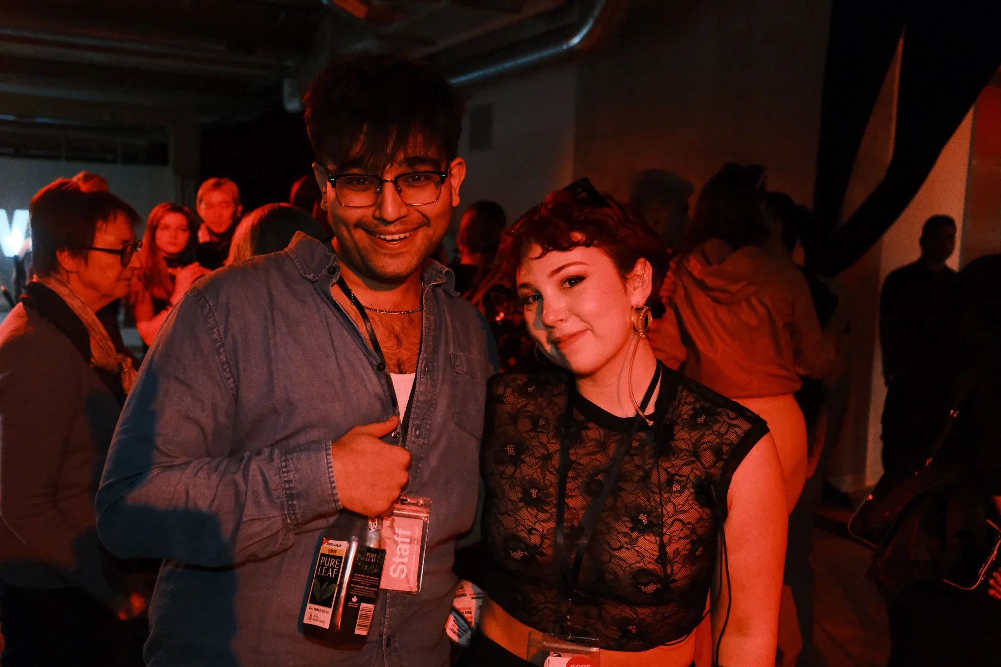 Two people smiling at a social event, with the man wearing glasses and a denim shirt, and the woman with earrings and a black lace top, surrounded by others in a dimly lit room.