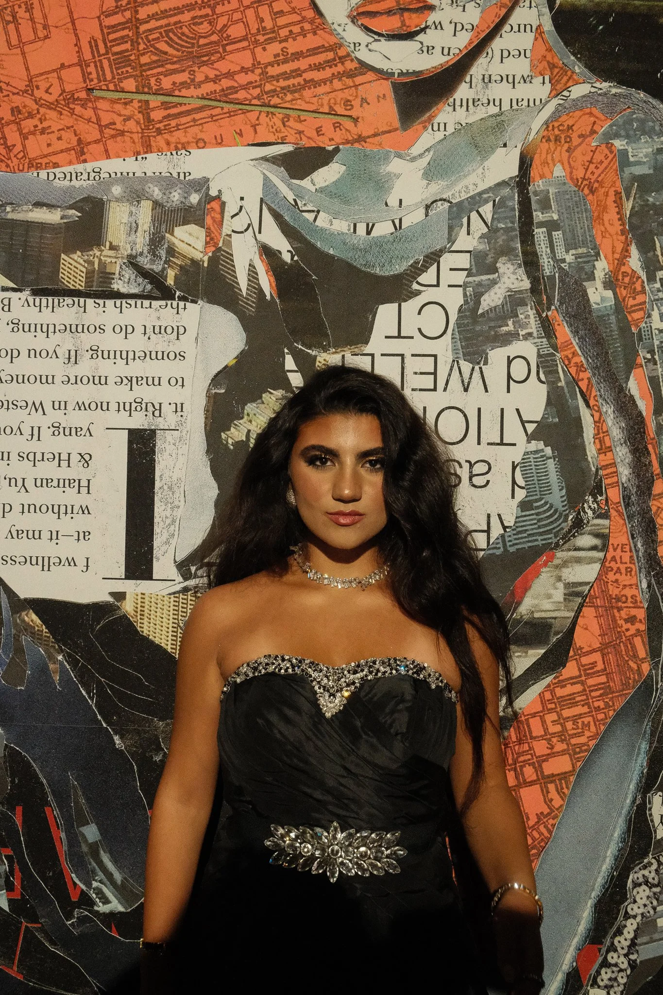A woman in a black strapless gown with silver embellishments and jewelry standing in front of a collage-style mural with newspaper clippings, maps, and images.