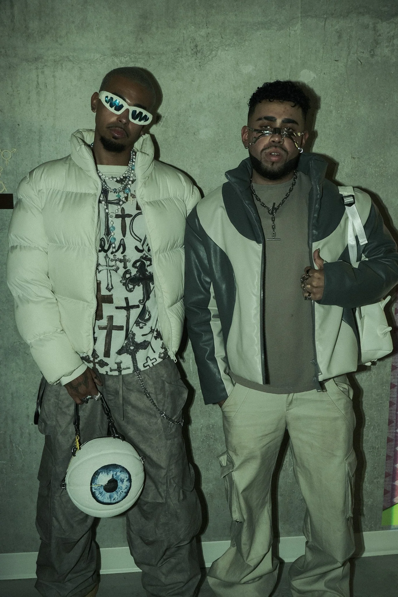 Two men standing against a gray wall, wearing stylish casual outfits, sunglasses, and jewelry, one holding a bag with an eye design.