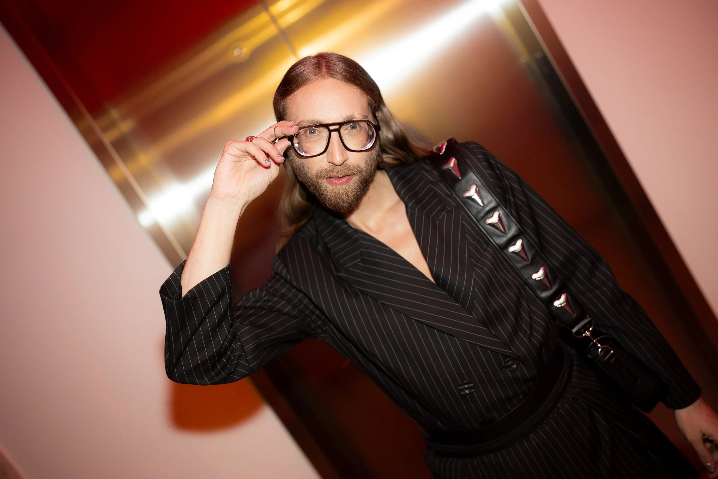 A man with long hair, a beard, and glasses, wearing a black pinstripe suit with metallic embellishments on his shoulder strap, adjusting his glasses while standing in an indoor setting with warm lighting.