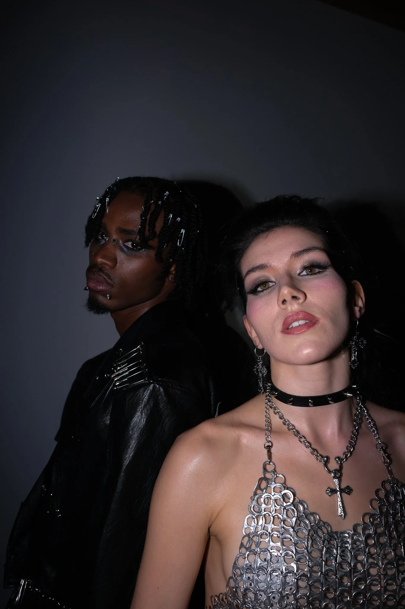 A man with black braided hair decorated with silver pins and a woman with dark hair styled back, wearing goth-style jewelry including a cross pendant, choker, and layered chains, looking at the camera against a dark background.