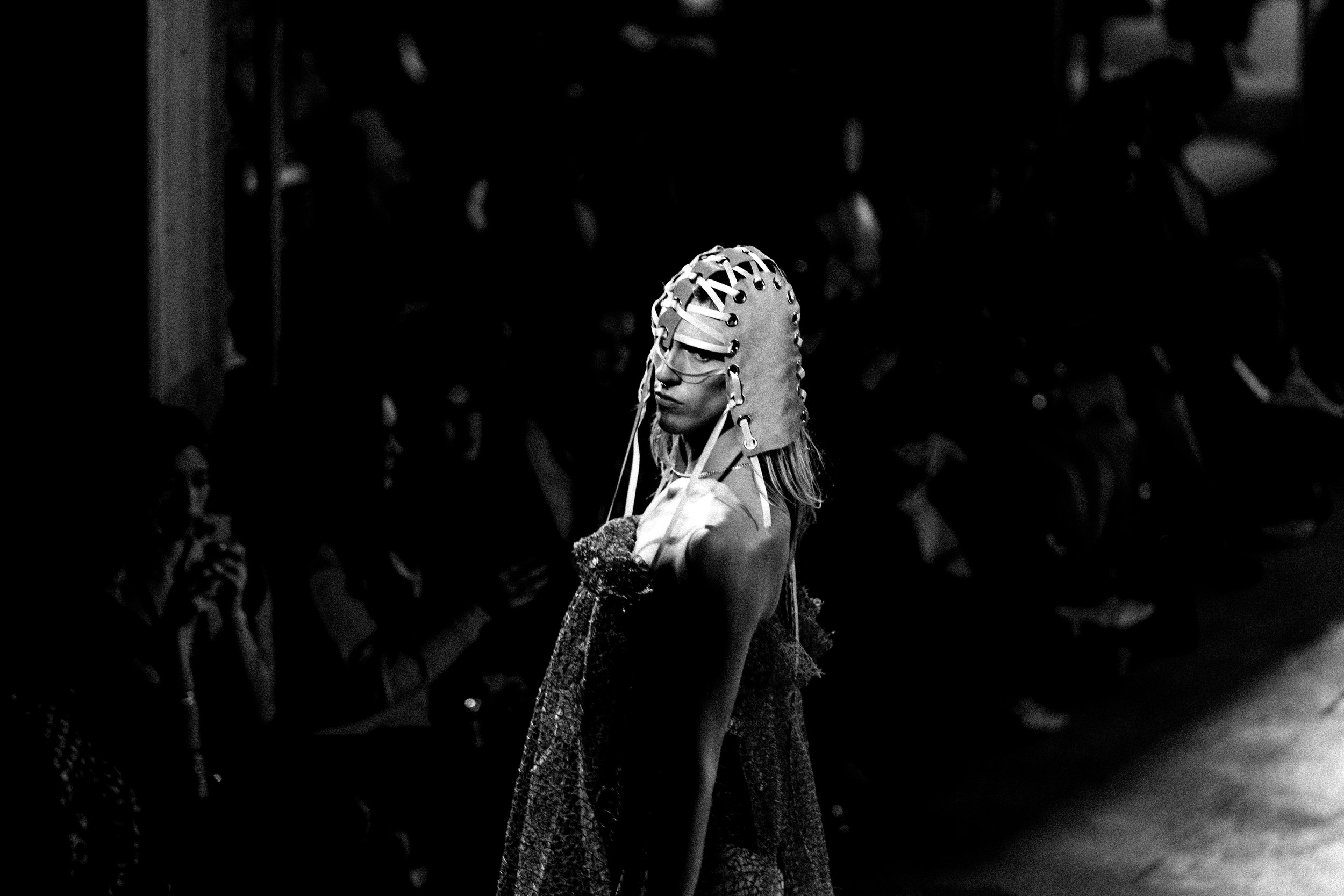 A model walking down a runway wearing a futuristic headpiece and a textured dress, with an audience watching in the background.