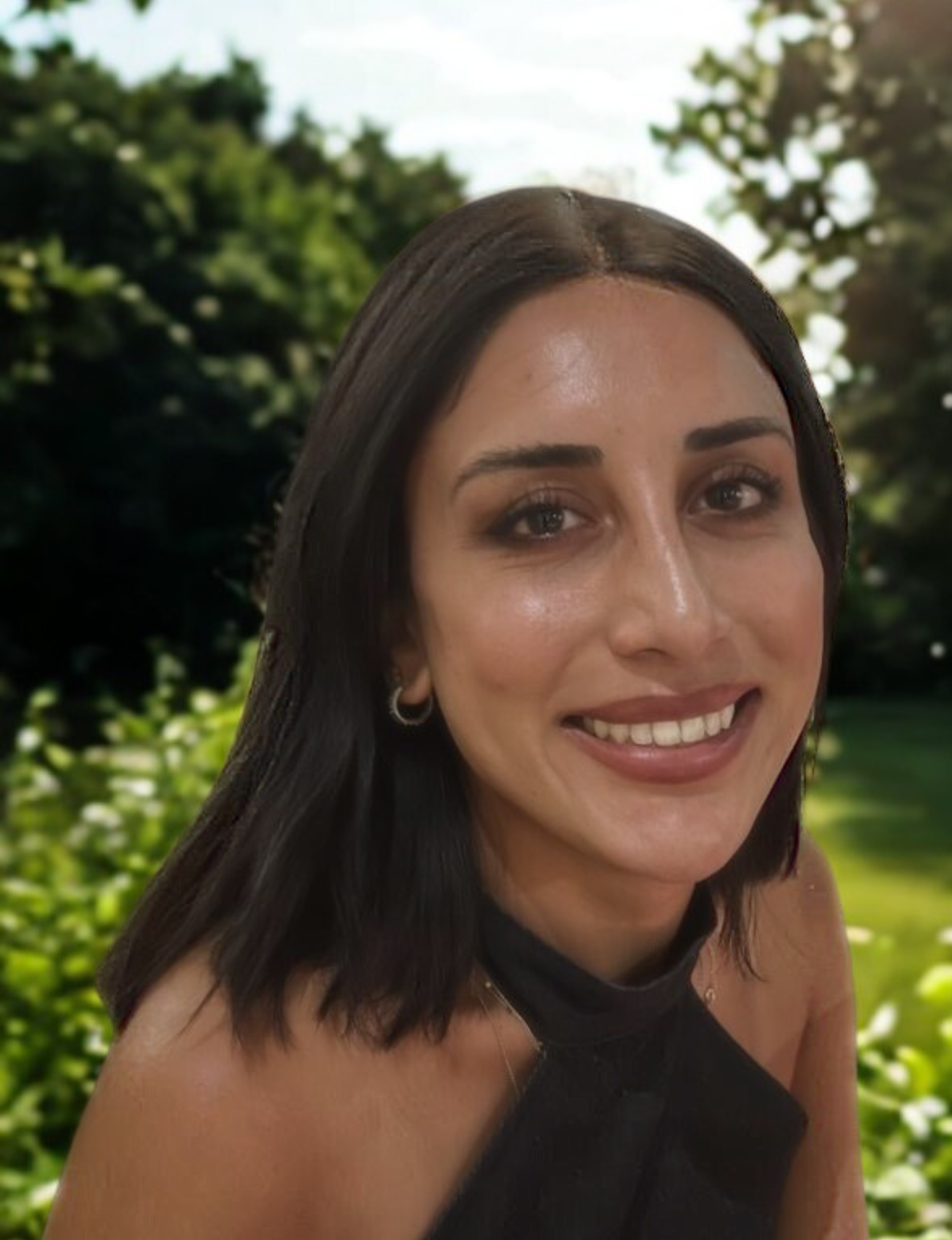 A woman with straight black hair, wearing hoop earrings and a black sleeveless top, smiling outdoors with green trees in the background.