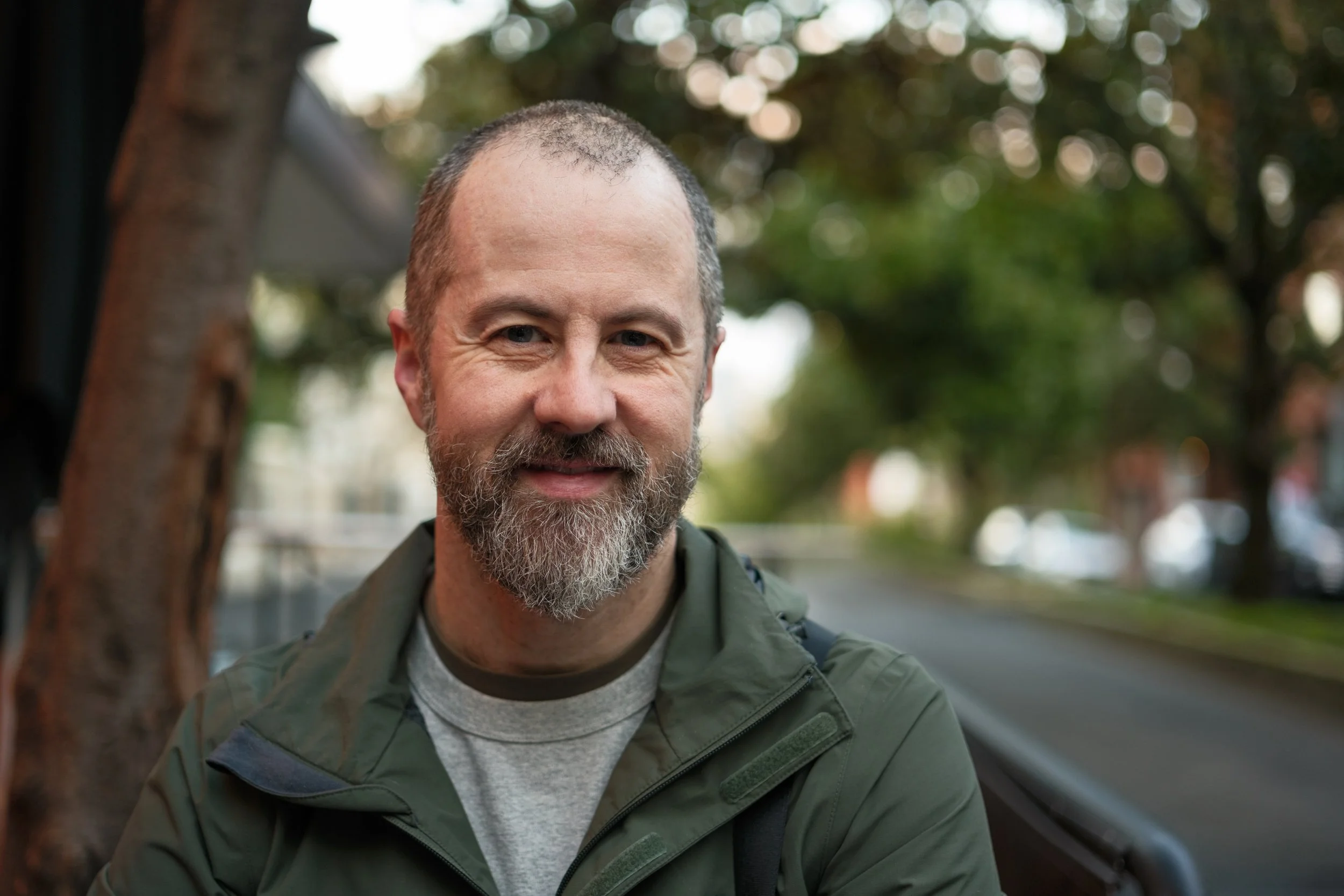 A smiling middle-aged man with a beard and short hair outside, wearing a green jacket and a gray shirt, sitting on a park bench near a tree with blurry trees and cars in the background.