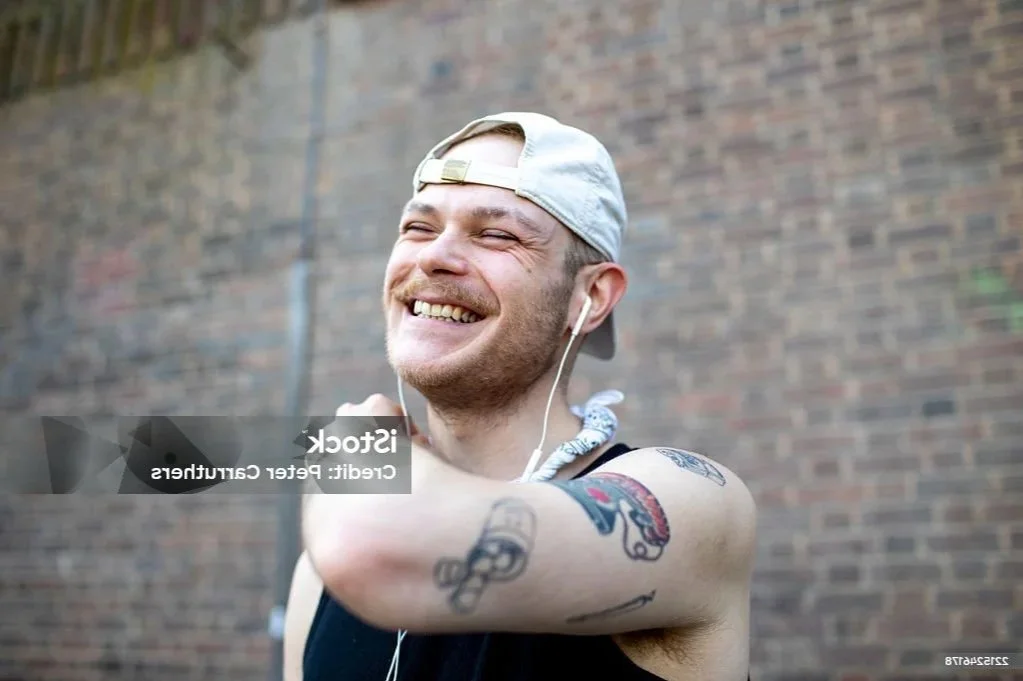 A smiling man with a baseball cap backwards, wearing a sleeveless shirt and earphones, standing outdoors against a brick wall.