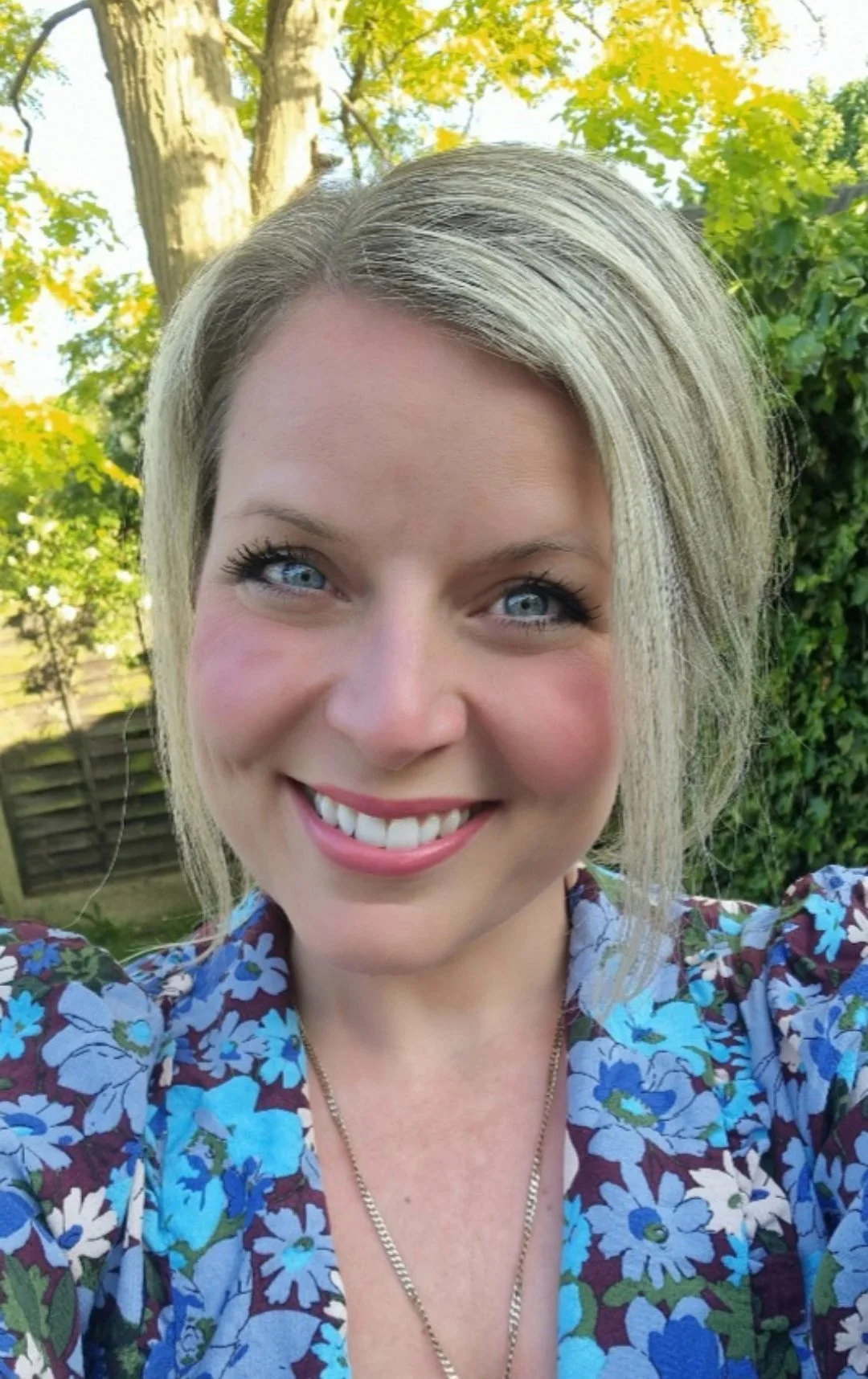A woman outdoors, smiling, with green trees in the background.