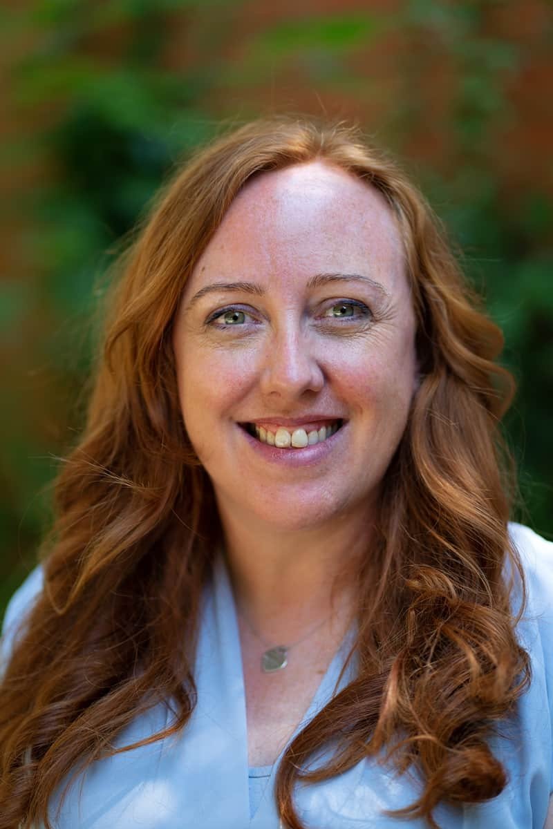 A woman with long, wavy red hair smiling outdoors, wearing a light blue top and a necklace, with a blurred green and brown background.