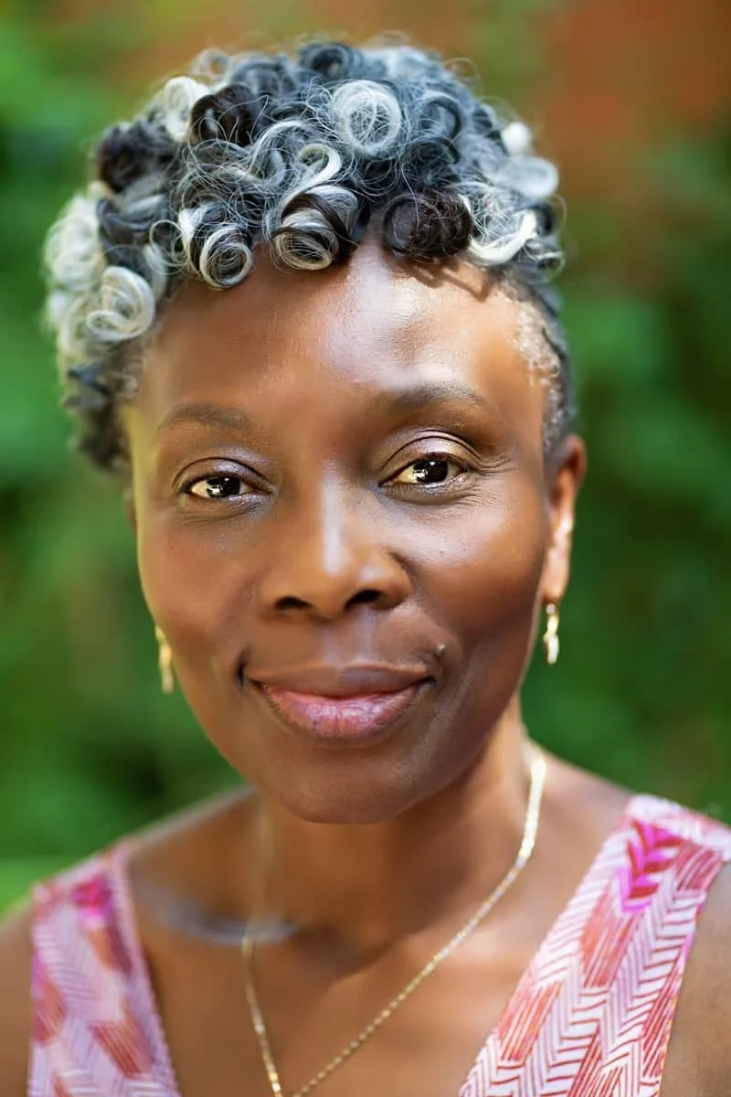 Close-up portrait of woman with short, curly hair with black and gray curls, smiling gently outdoors with a blurred green background, wearing a pink and white patterned sleeveless top and gold jewelry.