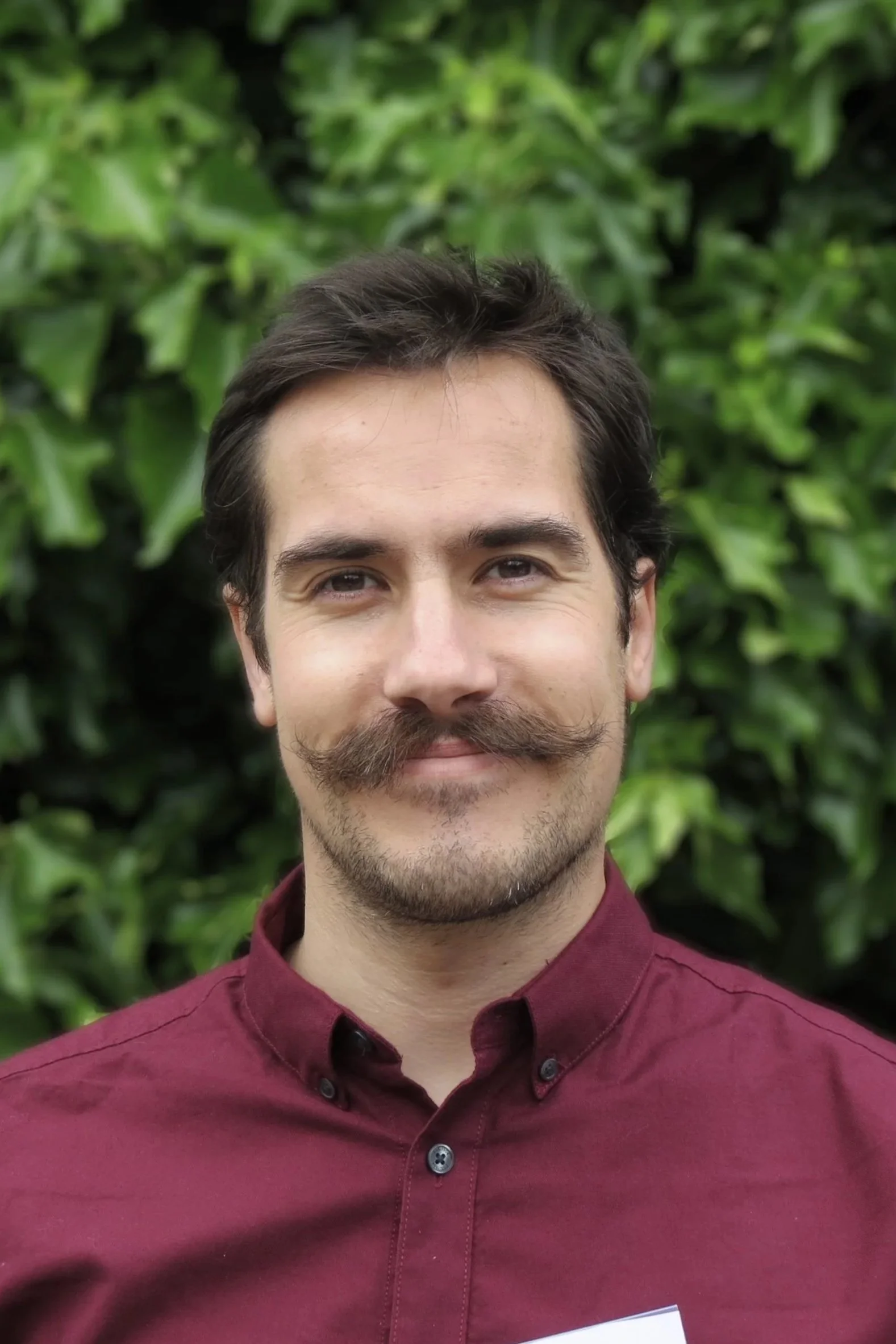 A man with dark hair, a mustache, and a beard, wearing a maroon shirt, standing outdoors in front of green foliage.