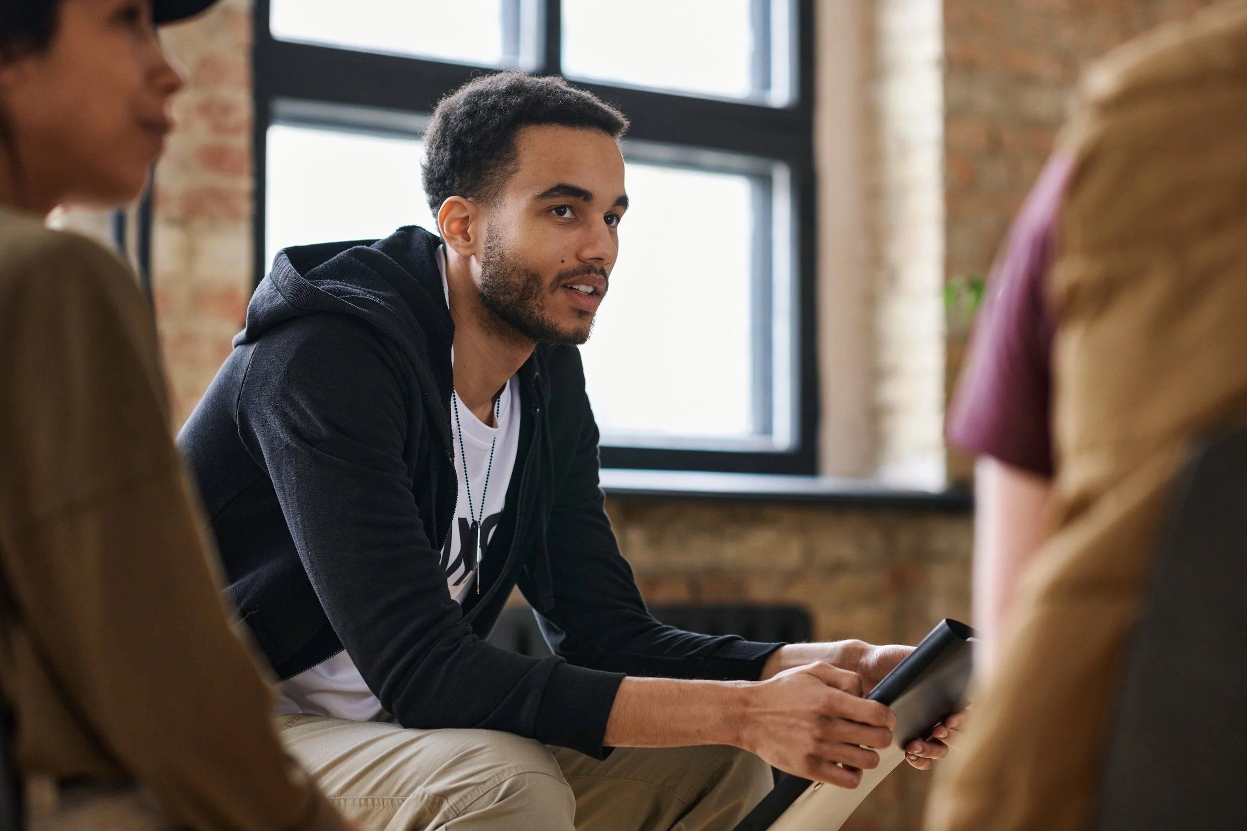 Young man talking in a group discussion, holding a tablet, with two women also participating.