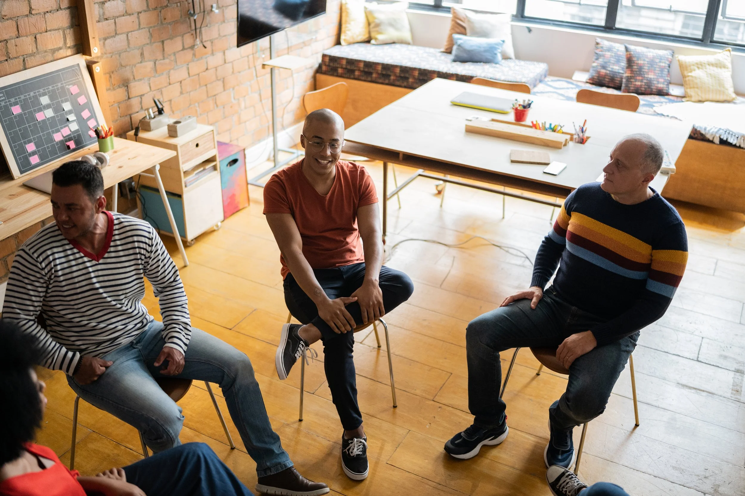 Four people sitting in a circle having a discussion in an office with a brick wall, large windows, and a long table in the background.