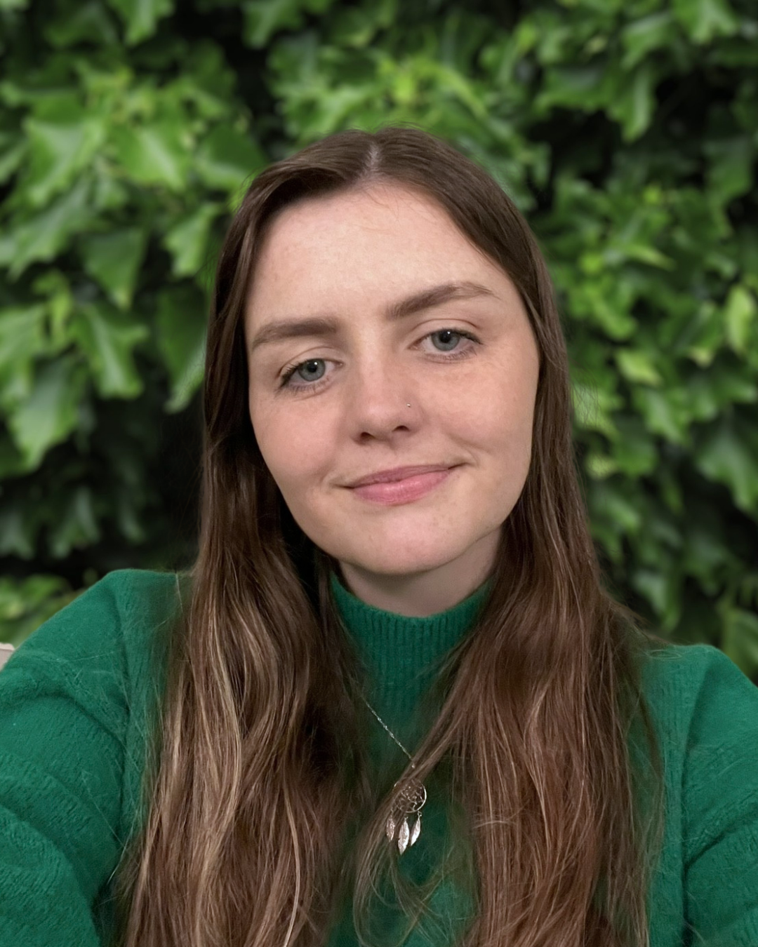 A woman with long brown hair and blue eyes, wearing a green sweater and a silver necklace, sitting in front of a leafy green background.