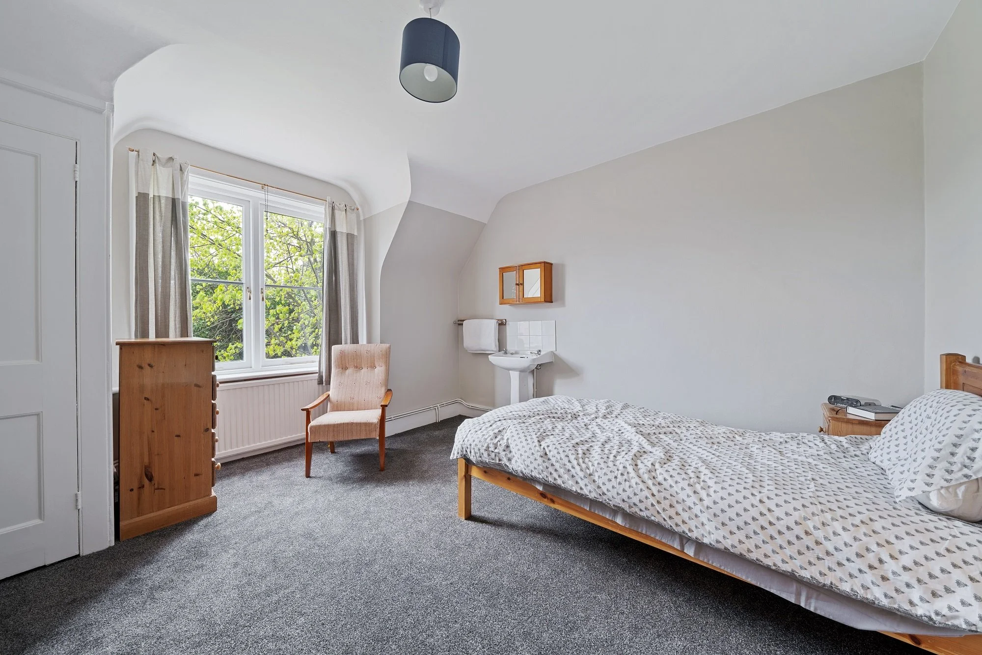 A simple bedroom with a single bed, wooden dresser, armchair near window, and a small sink with a towel rack on the wall.