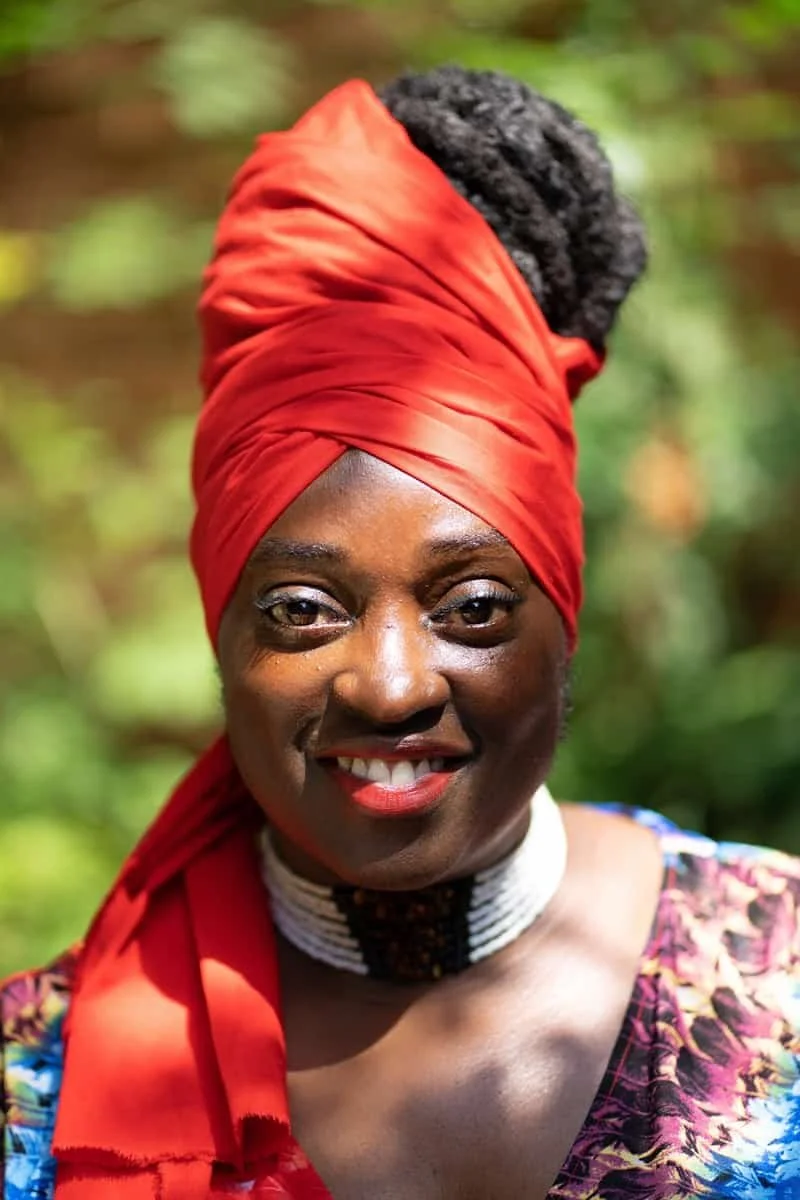 A woman smiling outdoors wearing a vibrant red headwrap, a patterned top, and a black and white beaded necklace.
