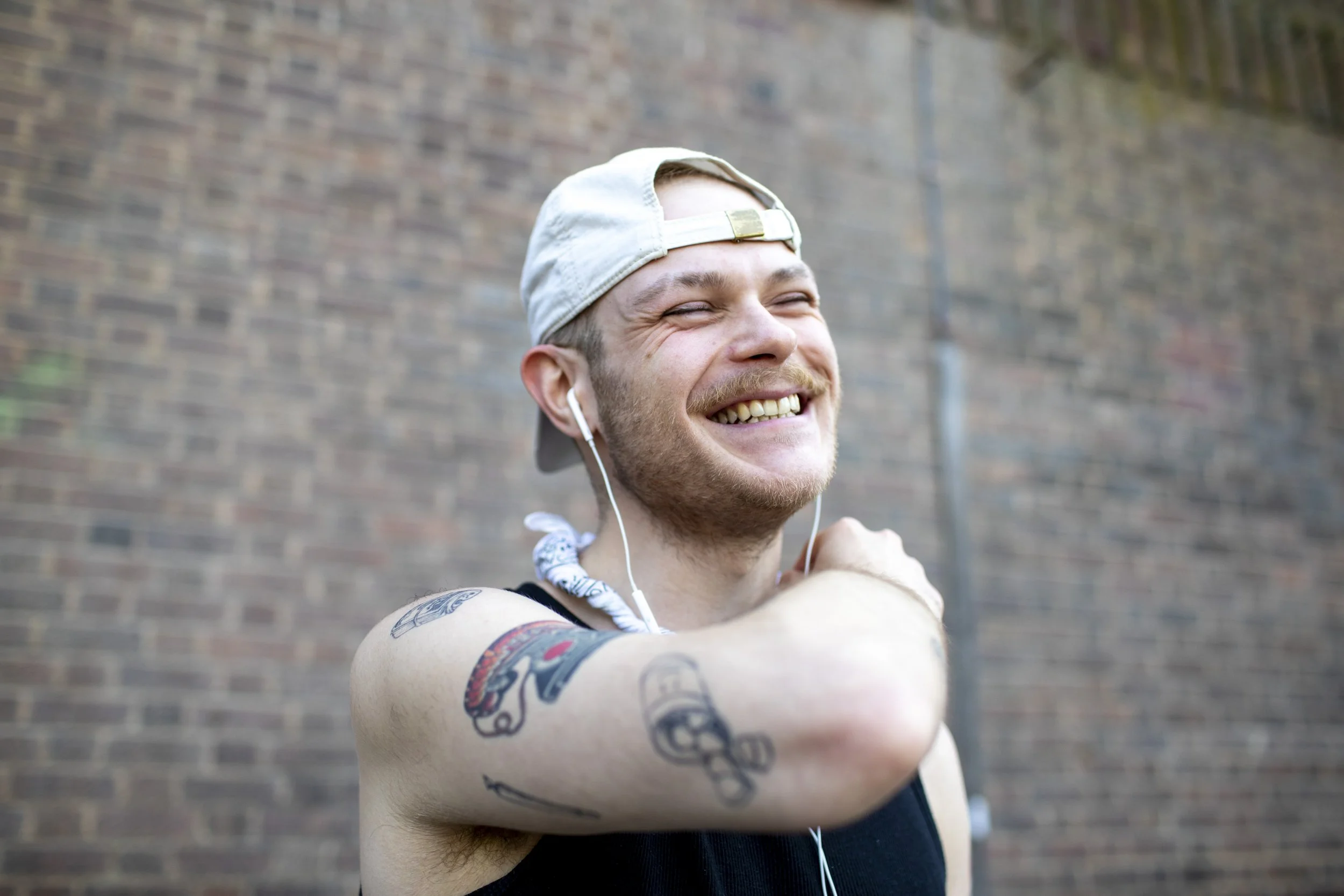 A smiling young man wearing a backwards cap, white earphones, and a sleeveless shirt, standing outdoors against a brick wall.