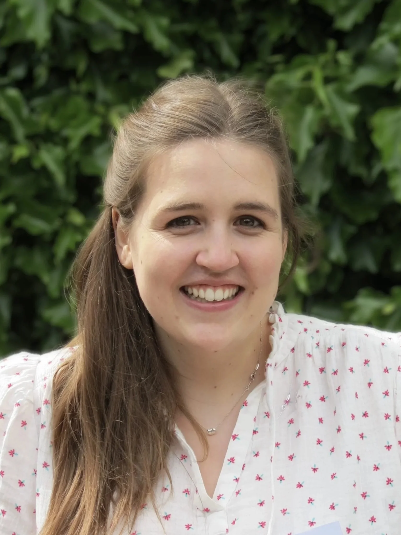 Close-up of a young woman with long brown hair, smiling, with greenery in the background, wearing a white blouse with small pink floral patterns and a delicate necklace.