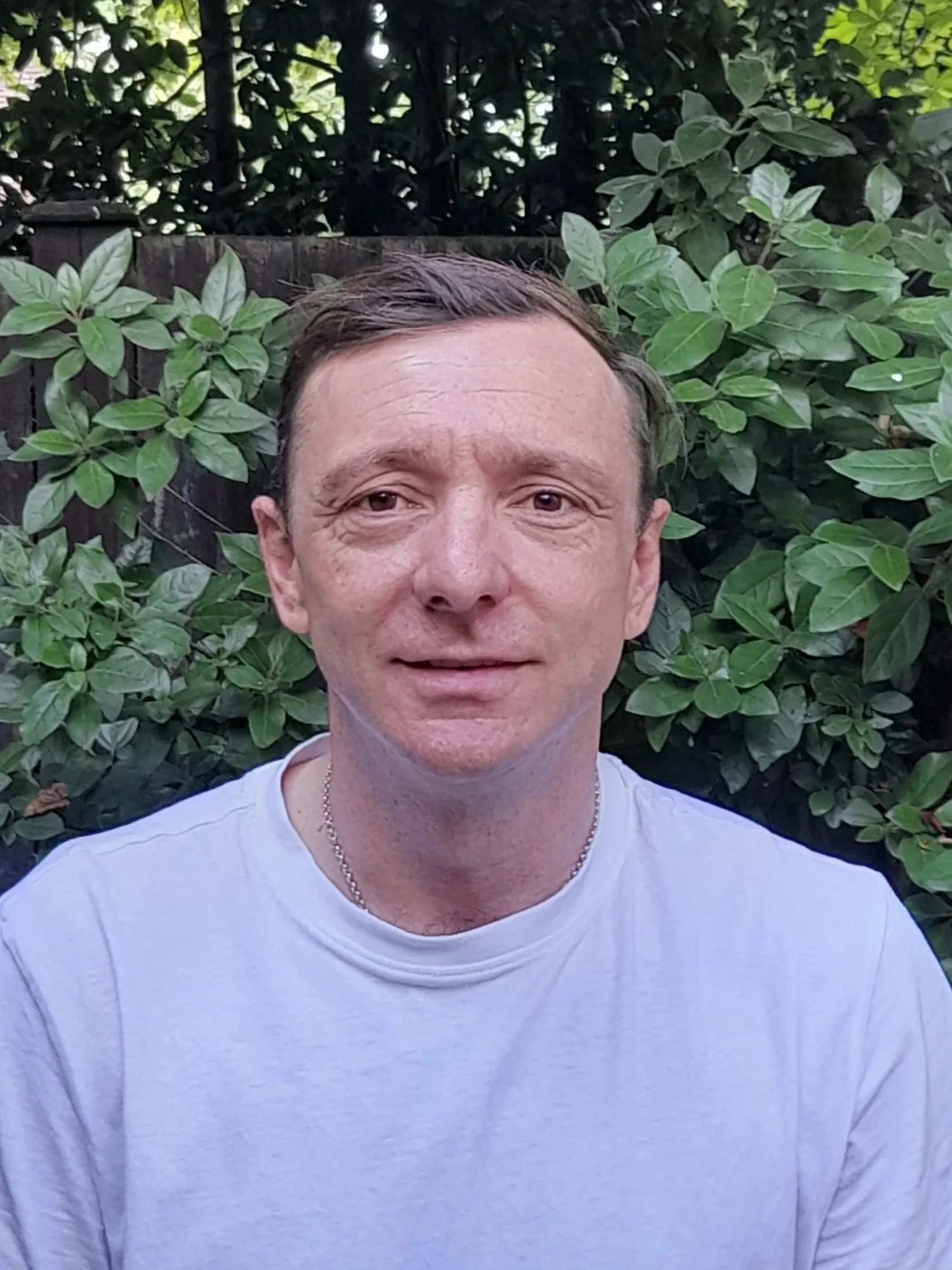 A man with brown hair and a white shirt is smiling while posing outdoors in front of green bushes and trees.