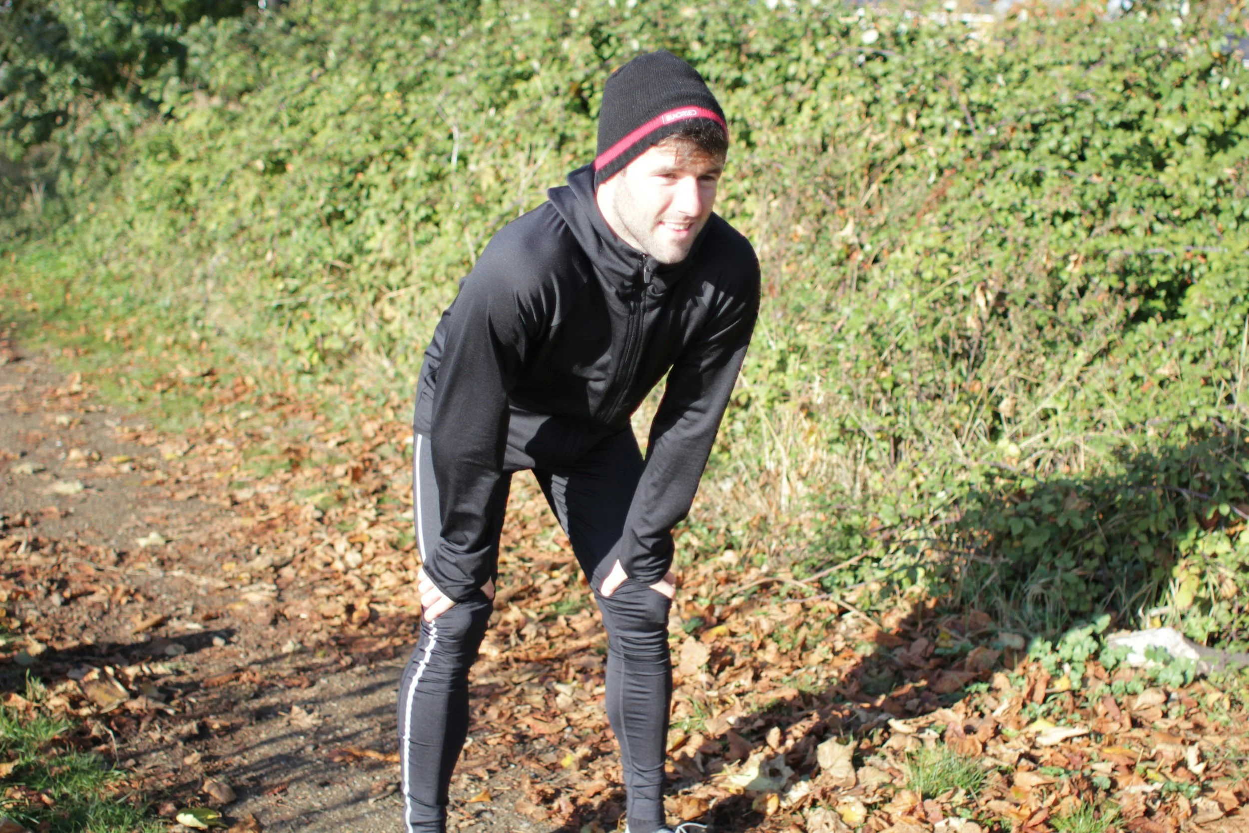 A young man dressed in black athletic wear, including a beanie, is outdoors on a forest trail surrounded by green bushes and fallen leaves, bending slightly forward with hands on his knees, possibly resting after exercising.