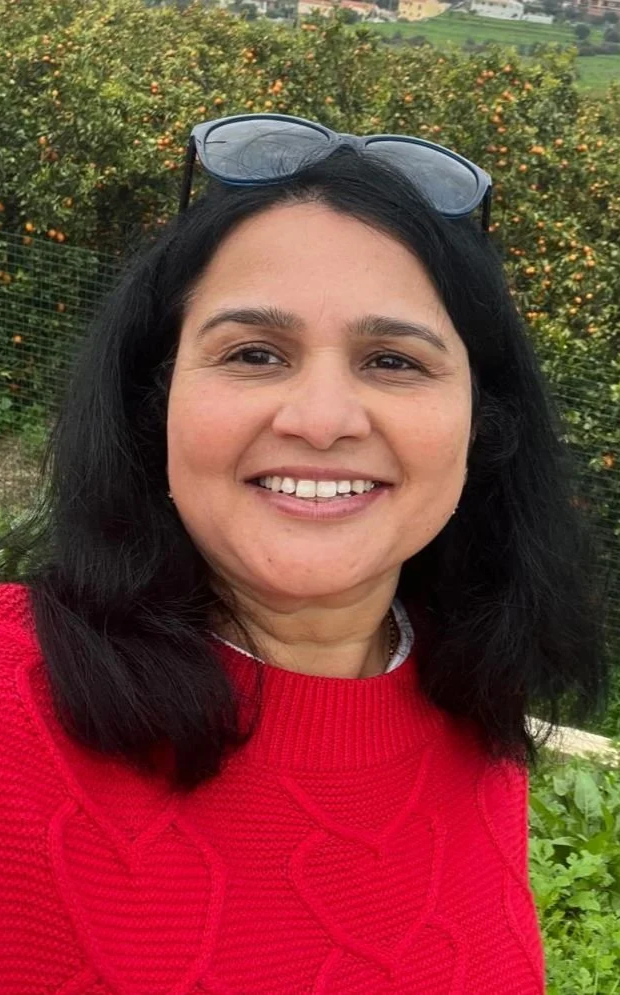 A woman with black hair smiling outdoors, wearing a red sweater and sunglasses on her head, with greenery and flowers in the background.