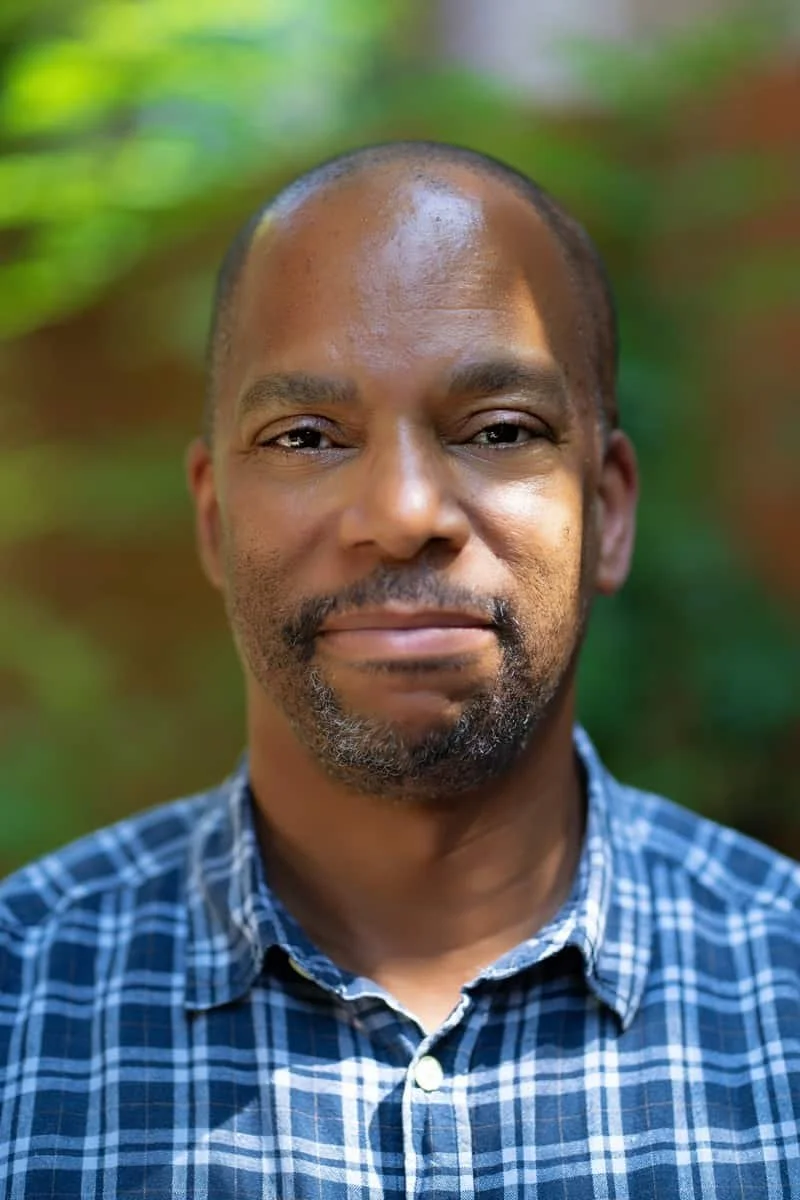 Close-up portrait of a man with a shaved head and goatee, wearing a blue plaid shirt, standing outdoors with blurred green background.