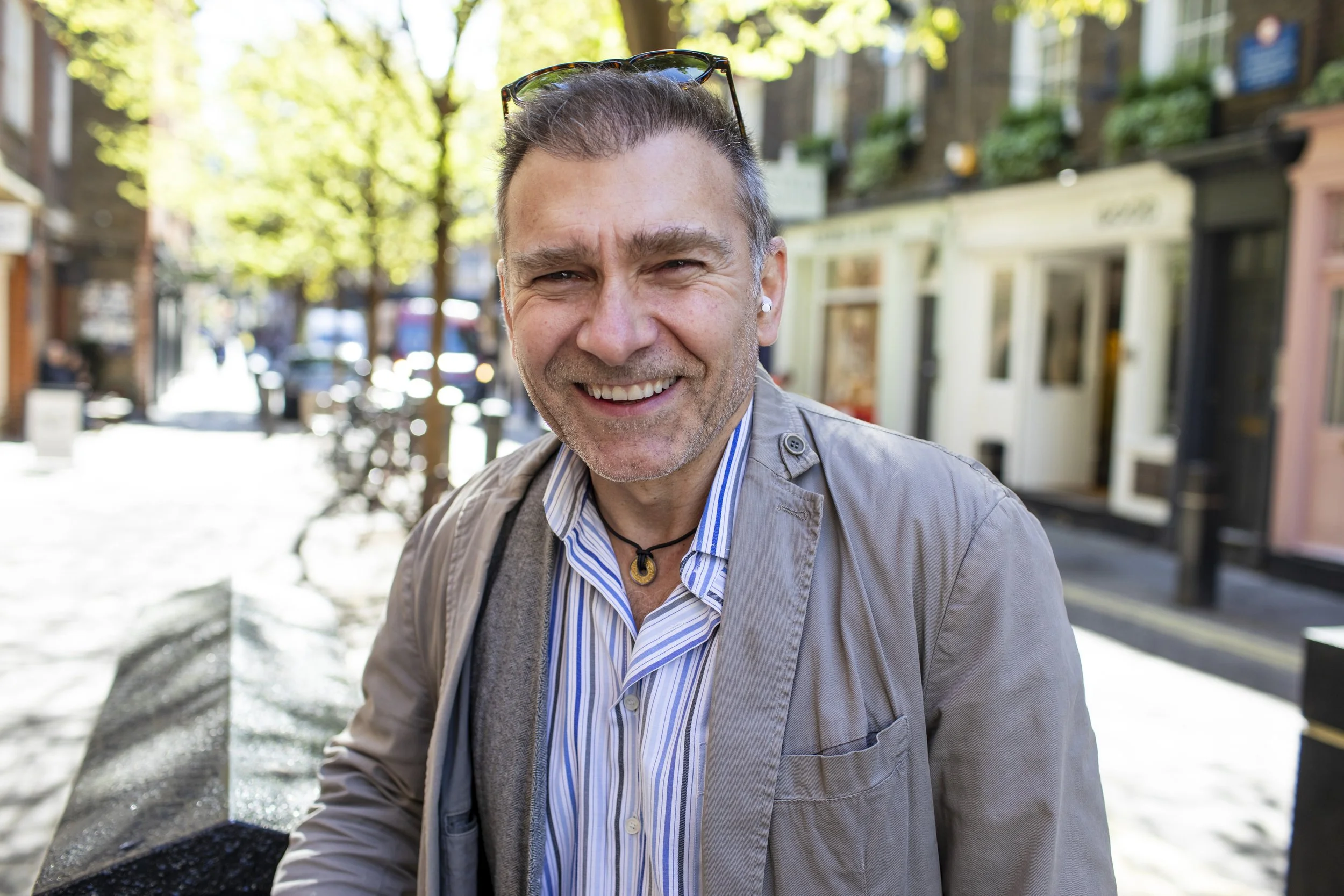 A smiling middle-aged man with sunglasses on his head, wearing a striped shirt and light gray jacket, standing outdoors on a sunny street with trees and storefronts in the background.