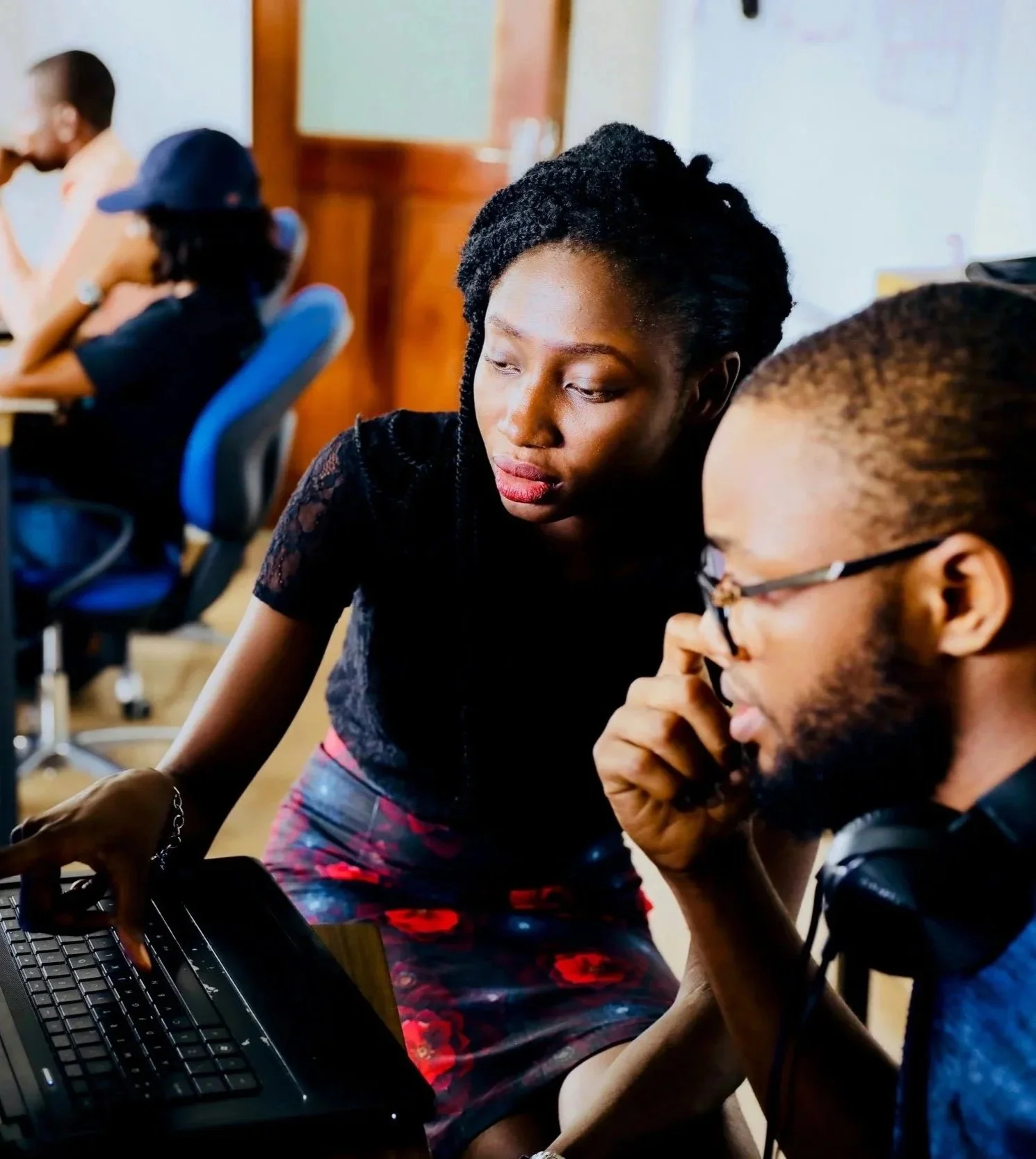 Two young adults, a woman with braided hair and a man with glasses and a beard, are collaborating on a laptop in a classroom or office setting, with other people working in the background.