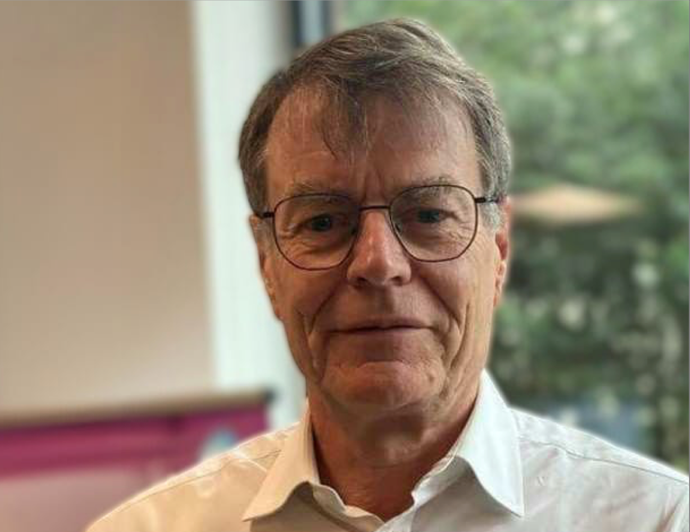 A man with glasses, short light brown hair, wearing a white shirt, standing indoors near a window with greenery outside.