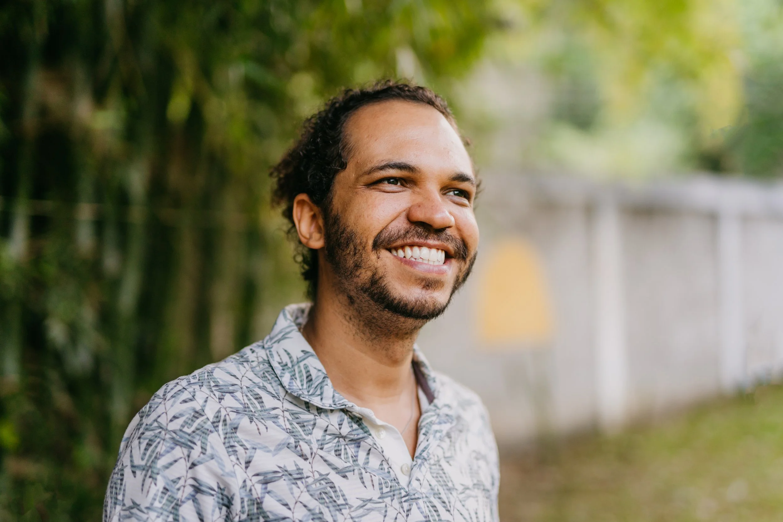 A smiling man with curly hair and a beard standing outdoors with trees and a blurred background.