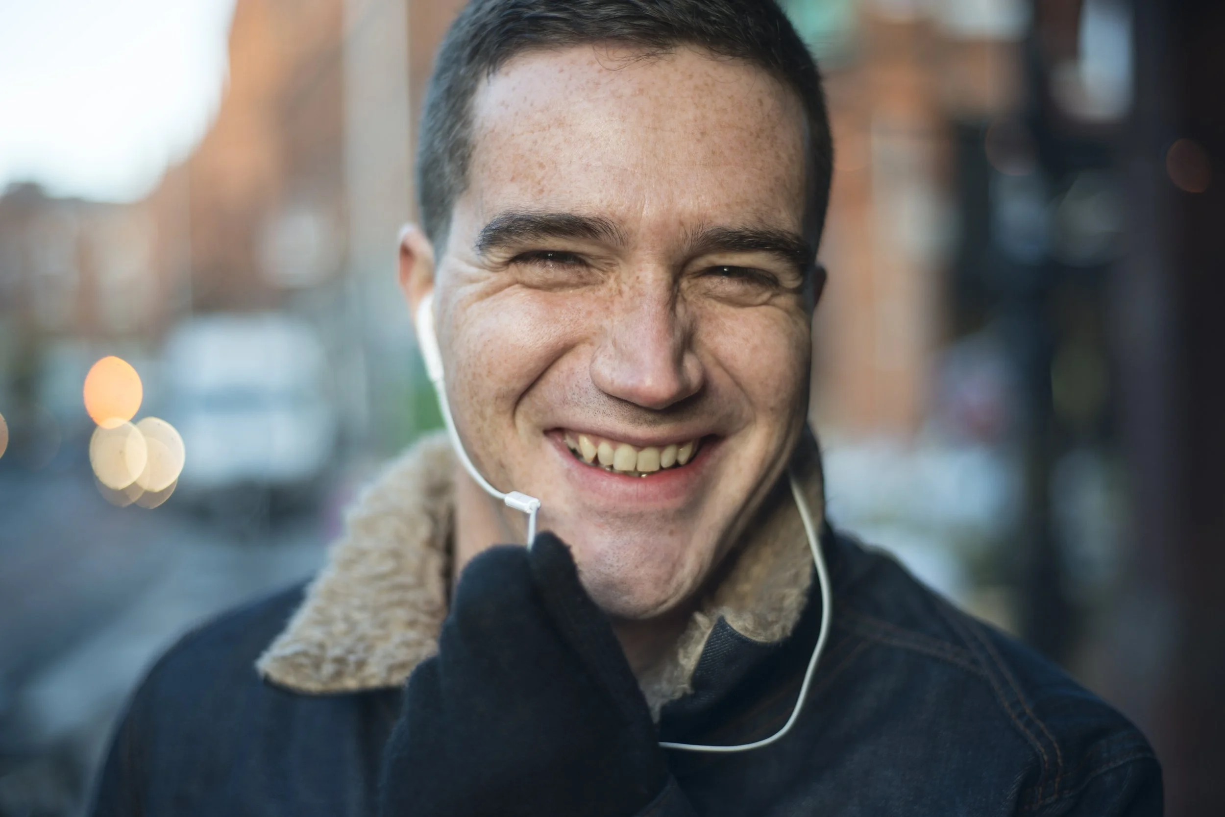 A young man smiling outdoors, wearing headphones and a jacket with a fleece collar, with blurred city street background.