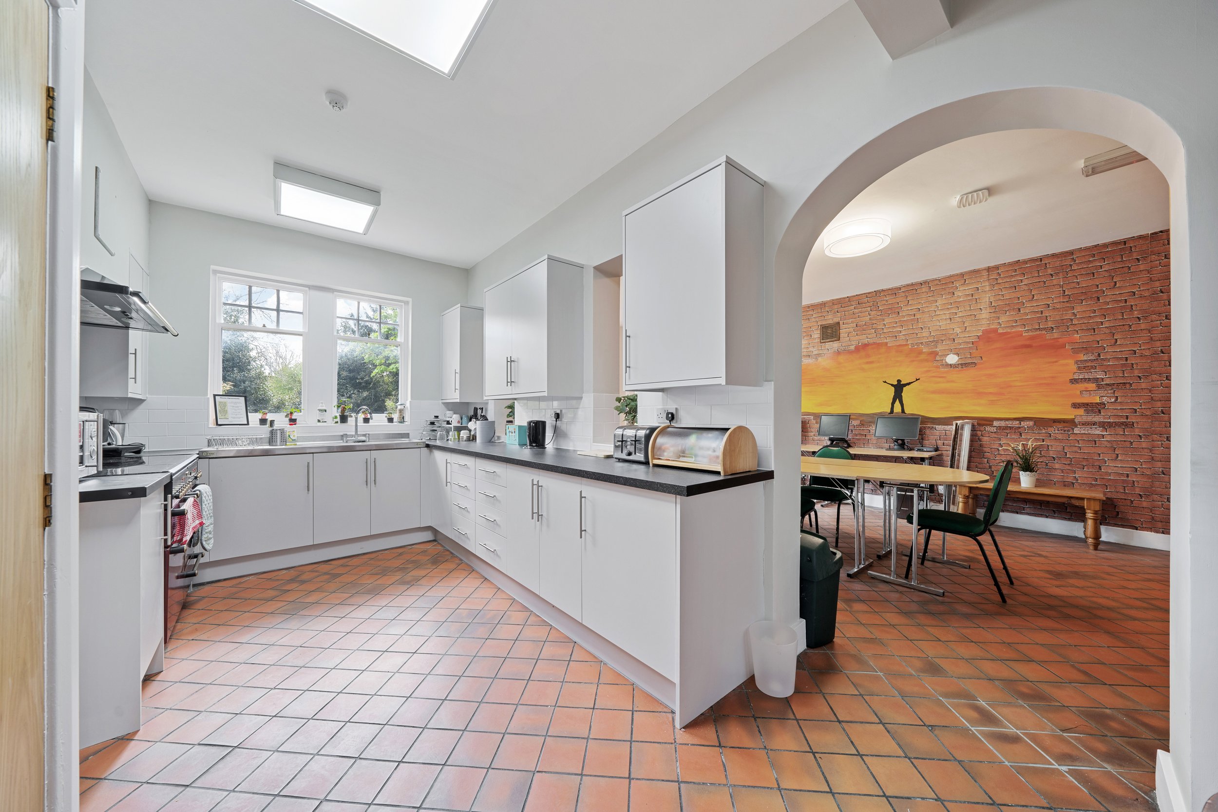 Kitchen with white cabinets, black countertop, terracotta tile floor, and a window showing outdoors; adjacent room with brick wall, desk, green chairs, and sunset mural.