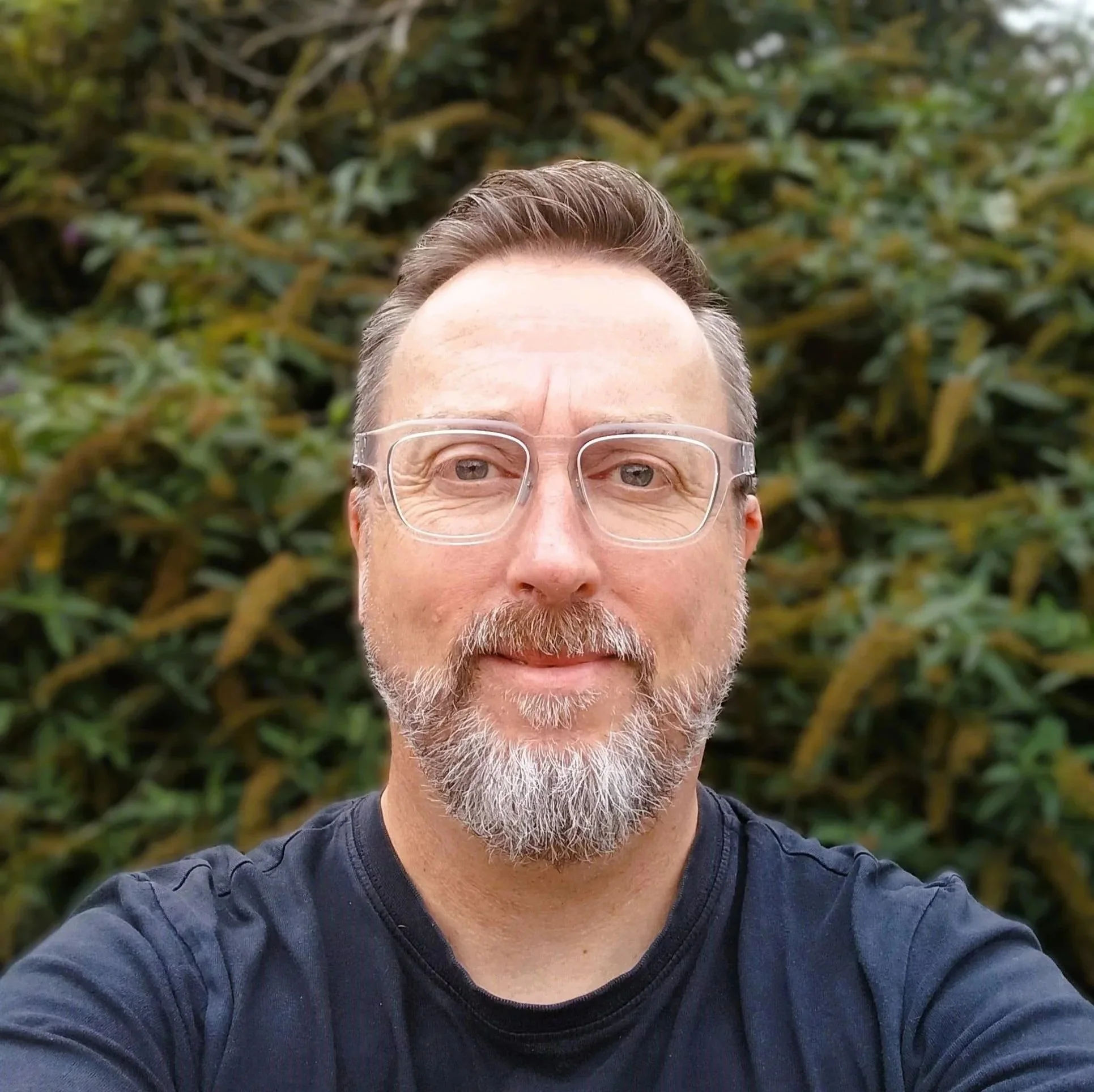 A man with glasses and a beard taking a selfie outdoors with a background of green foliage.